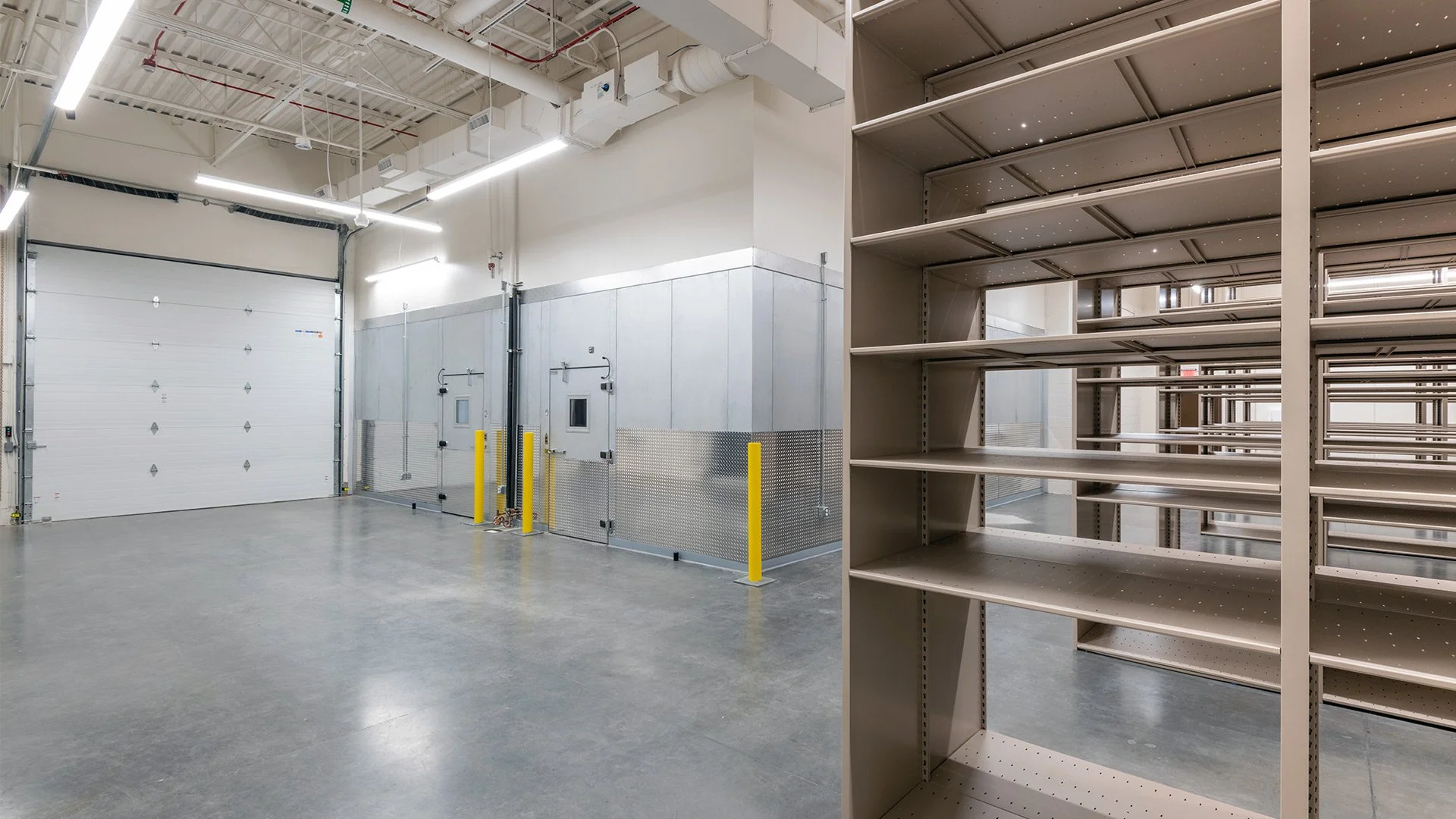 Empty warehouse with a large roll-up door, storage area with shelves, and some yellow protective barriers near metal doors on the wall.
