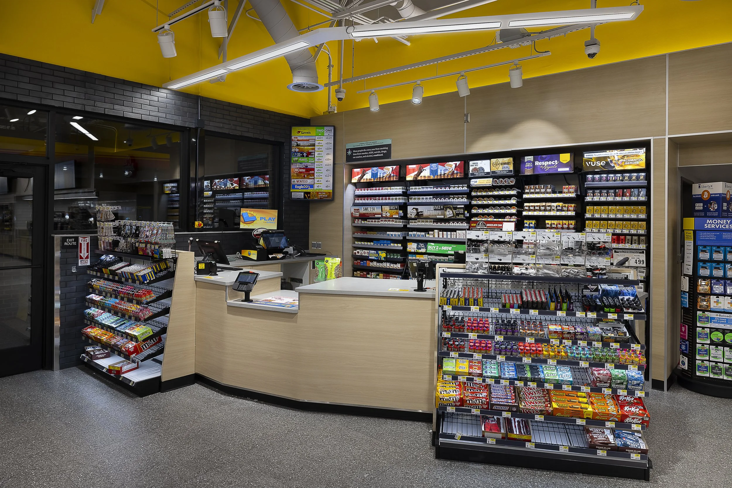 Inside a convenience store checkout area with candy and snacks on display, cash registers, digital screens, and a yellow ceiling.