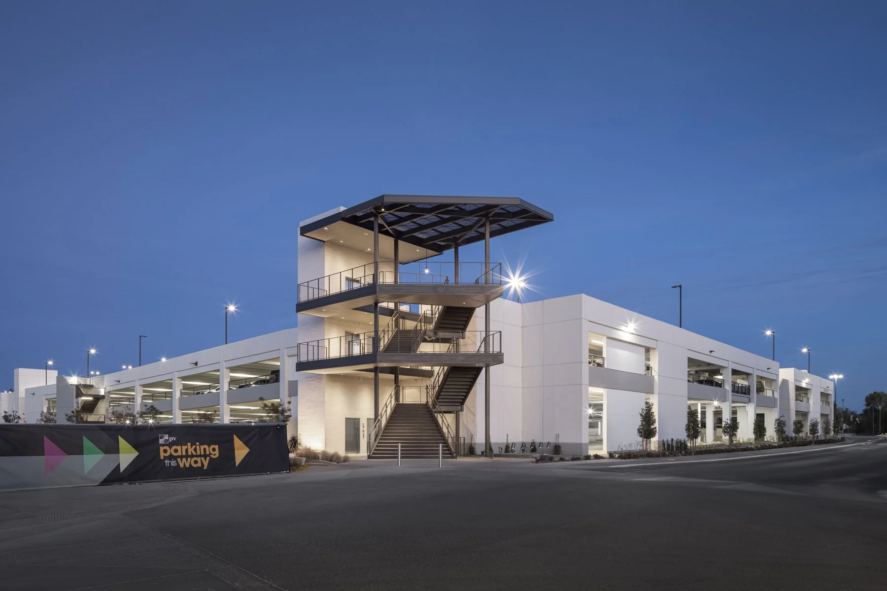 Modern multi-story parking garage building with exterior staircases and a rooftop terrace at dusk, illuminated by outdoor lighting.
