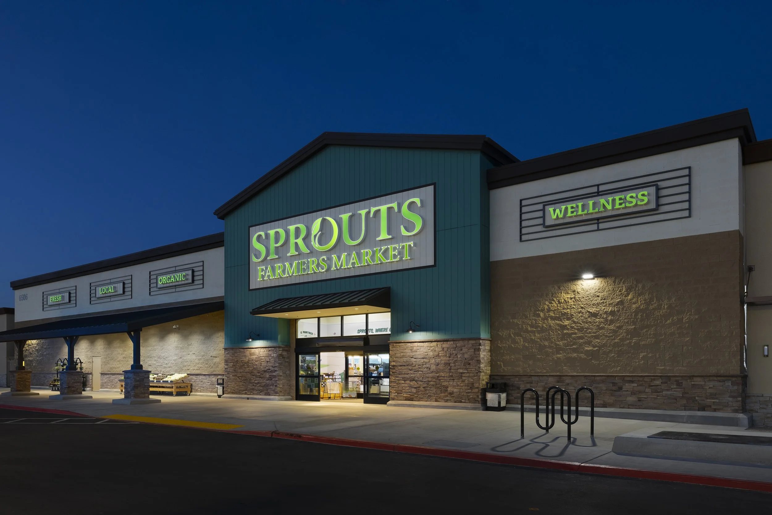 Exterior view of Sprouts Farmers Market grocery store at dusk with illuminated signs, including 'Sprouts Farmers Market' and 'Wellness'.