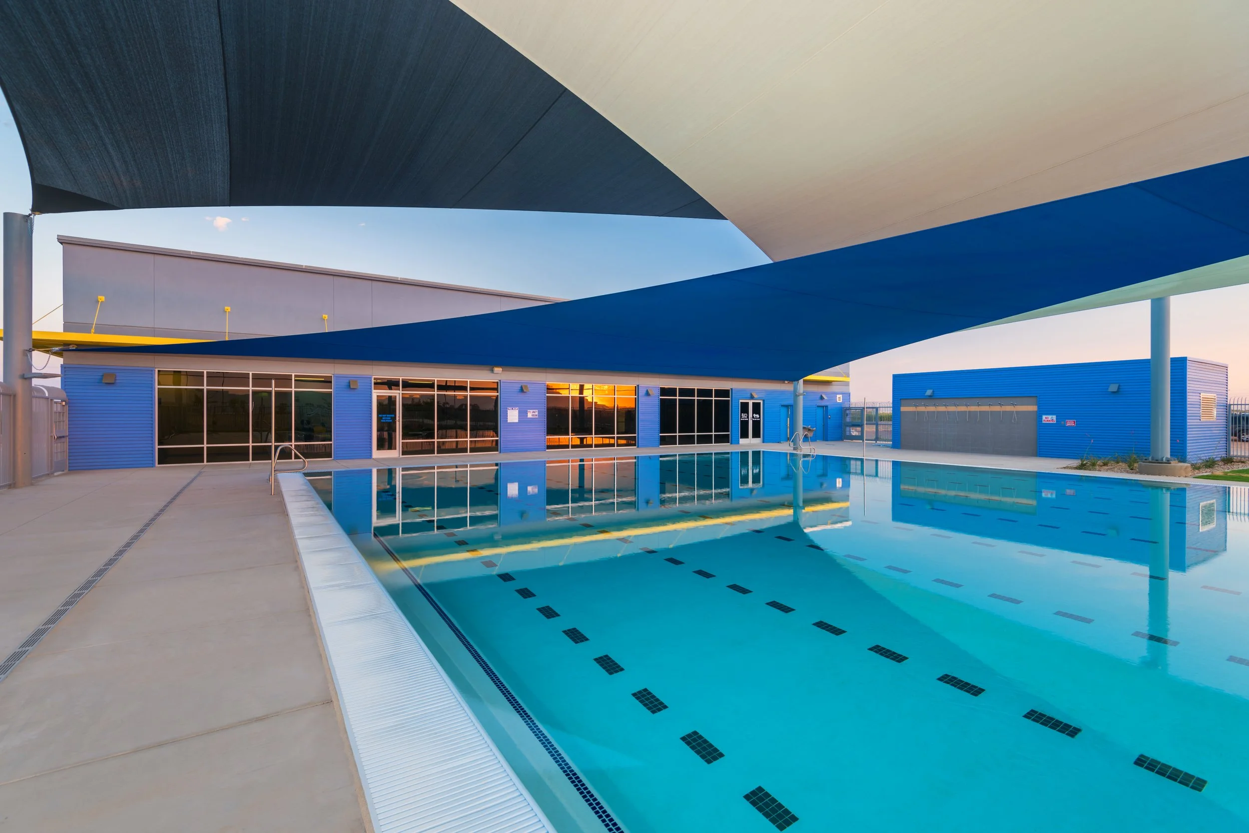 Empty outdoor swimming pool with blue shade sails and a modern blue building in the background, during sunset.
