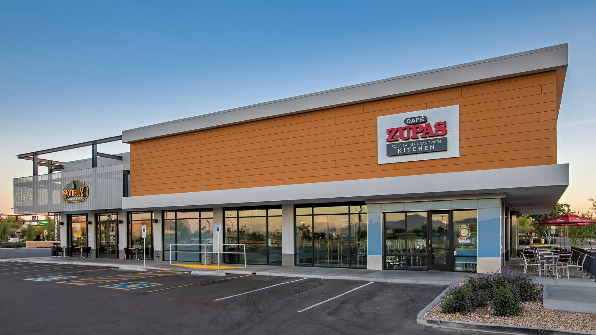 Exterior of a modern commercial building with signs for Potbelly sandwich shop and Cafe Zupas, featuring large windows, parking spots, and outdoor seating.