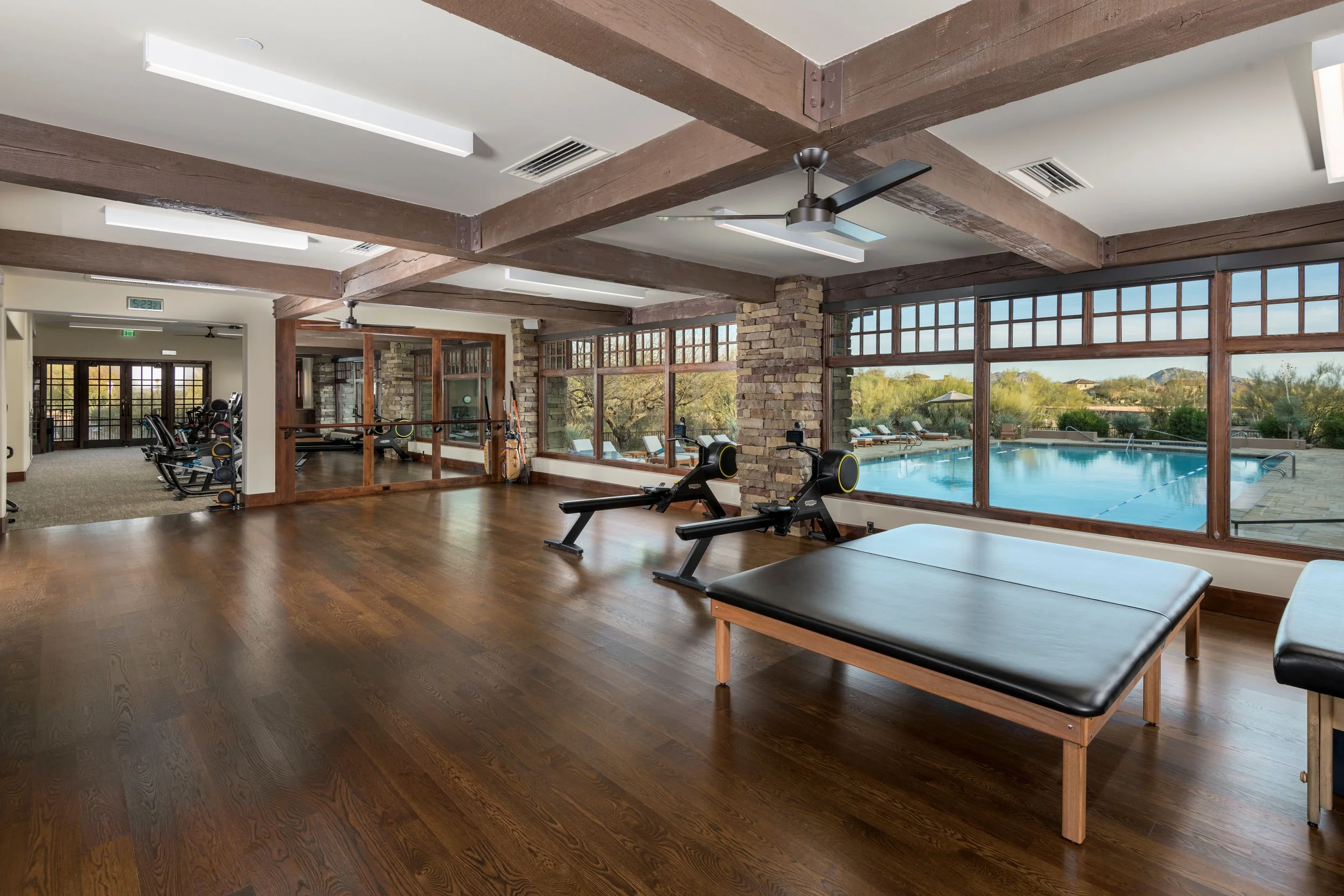 A spacious indoor gym with wooden floors, large windows showing outdoor pool area, exercise equipment including rowing machines, weight rack, and lounge chairs outside near the pool.