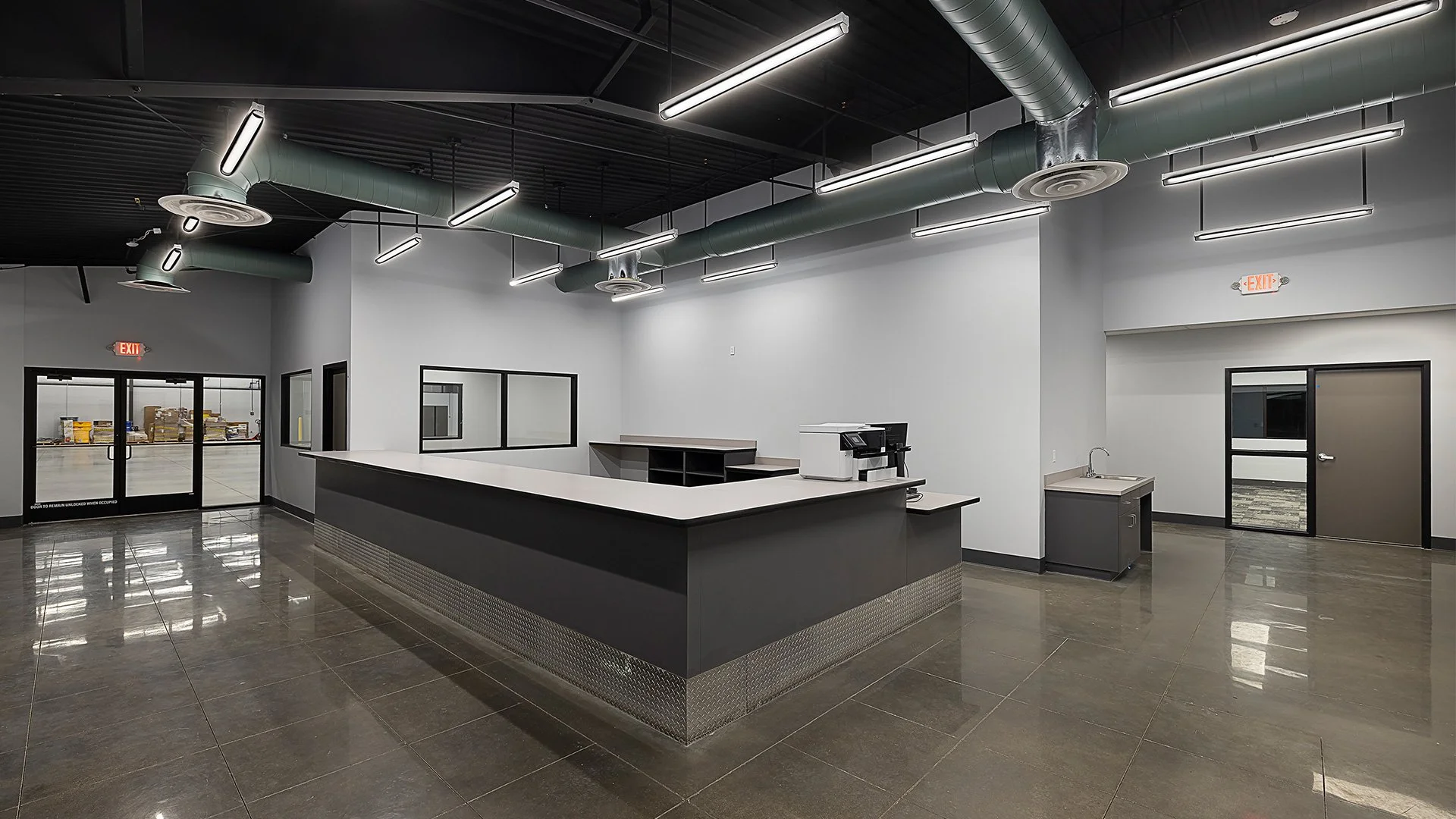 Empty commercial reception area with a black and white counter, coffee machine, sink, large windows, and doors to a warehouse or storage area. Exposed ceiling with ventilation ducts and modern lighting.