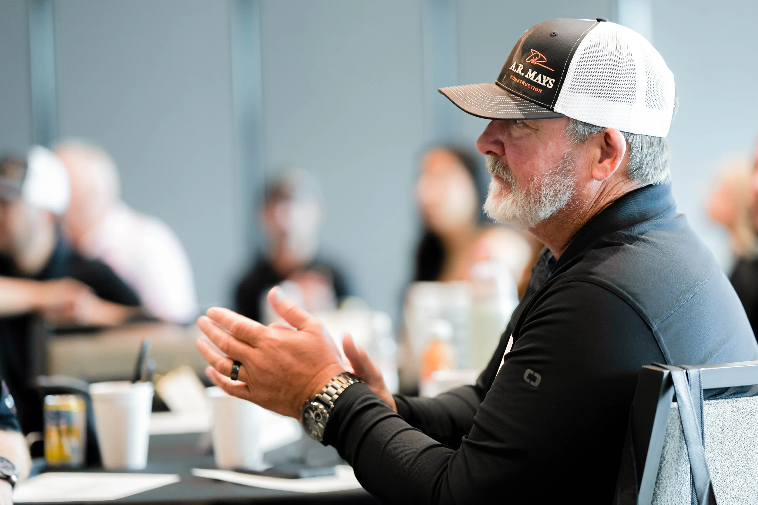 A man with a white beard wearing a black baseball cap and black long sleeve shirt sits at a conference table, clapping his hands, in a meeting room with blurred people in the background.