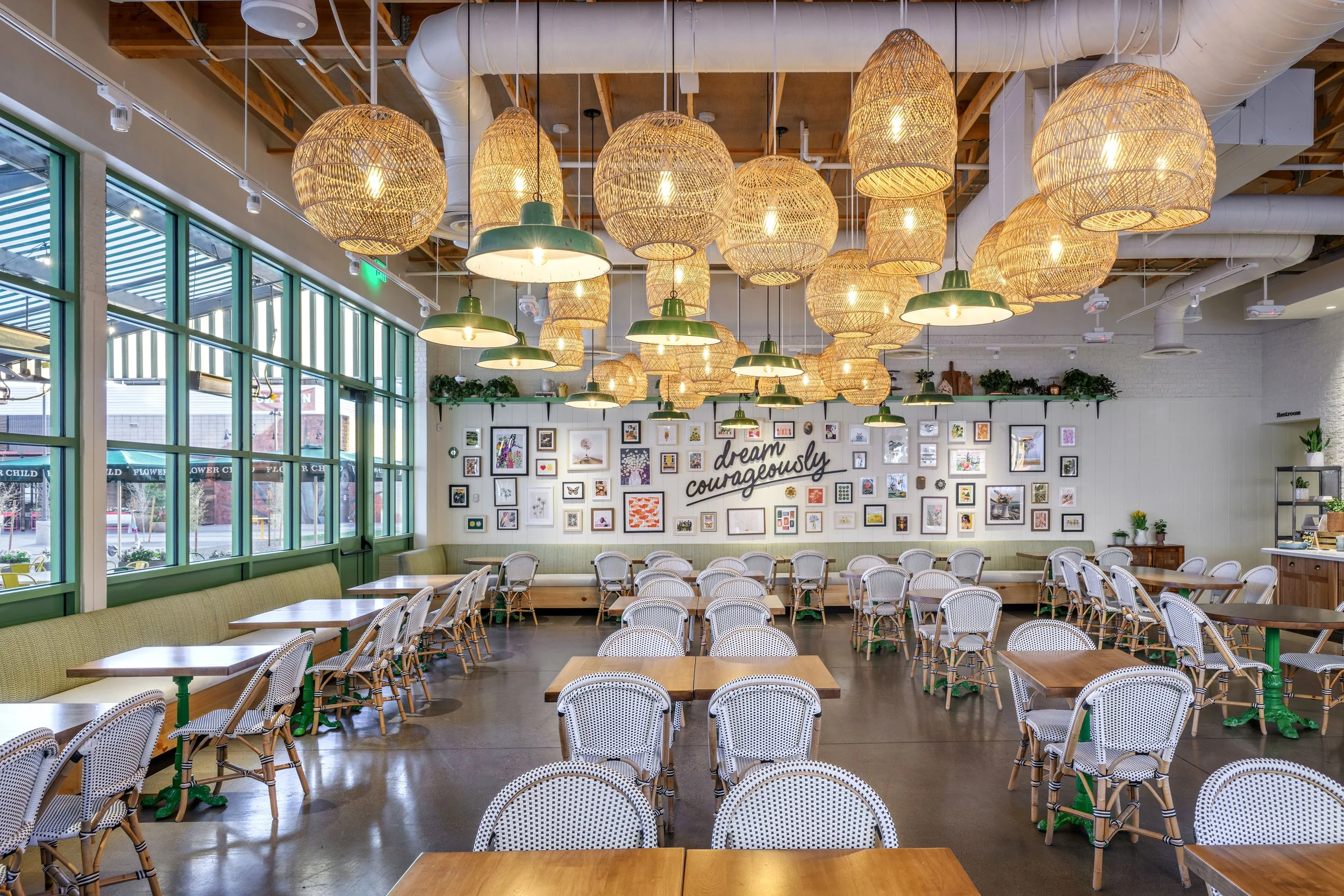 Interior of a bright, modern cafe with wooden tables and white wicker chairs, large windows, hanging woven light fixtures, and a white wall decorated with framed artwork and a sign that says 'dream courageously'.