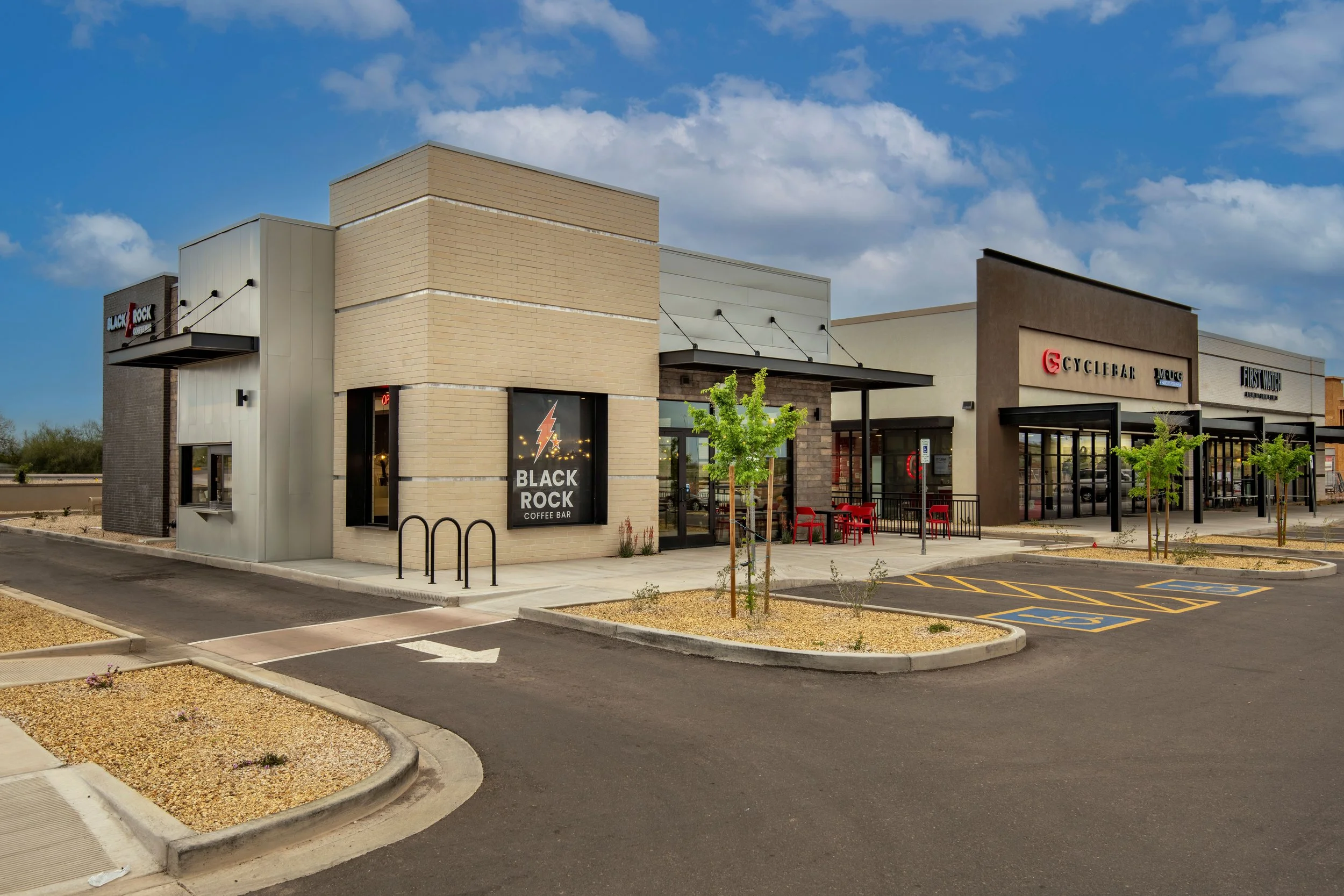 Exterior view of a shopping plaza with several storefronts, including Black Rock Coffee Bar, CycleBar, and First Watch, with a parking lot and a few small trees in the foreground.
