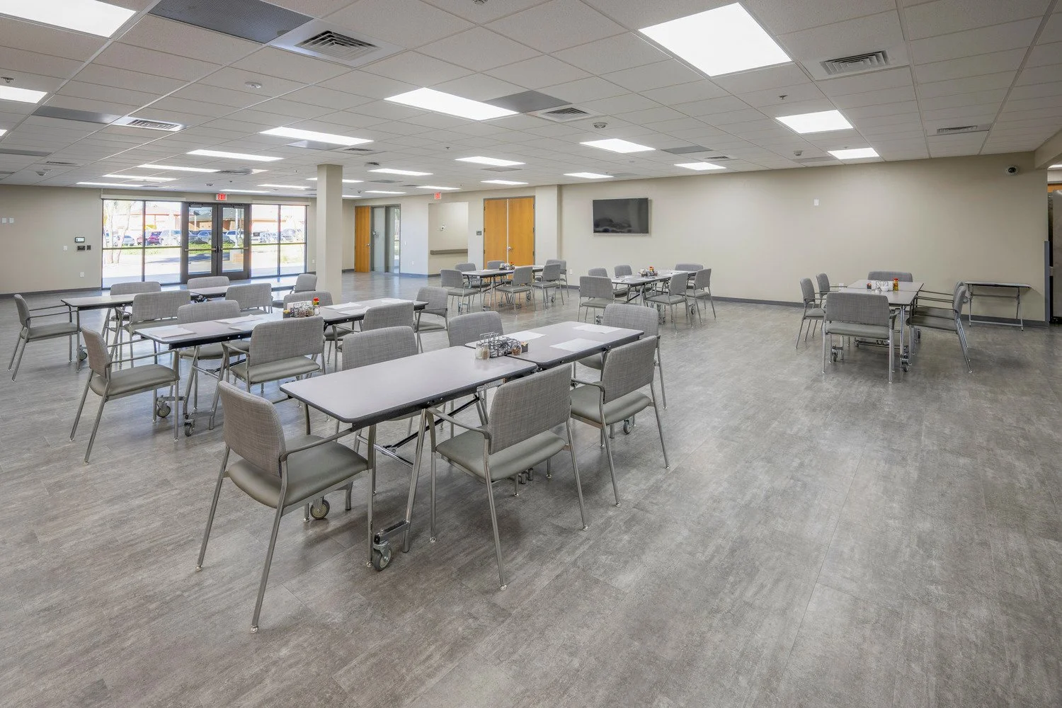 Empty dining or meeting room with multiple rectangular tables and gray upholstered chairs. Large windows at the front with glass doors, and a wall-mounted television on the right. Neutral-colored walls and gray flooring, with overhead fluorescent lig