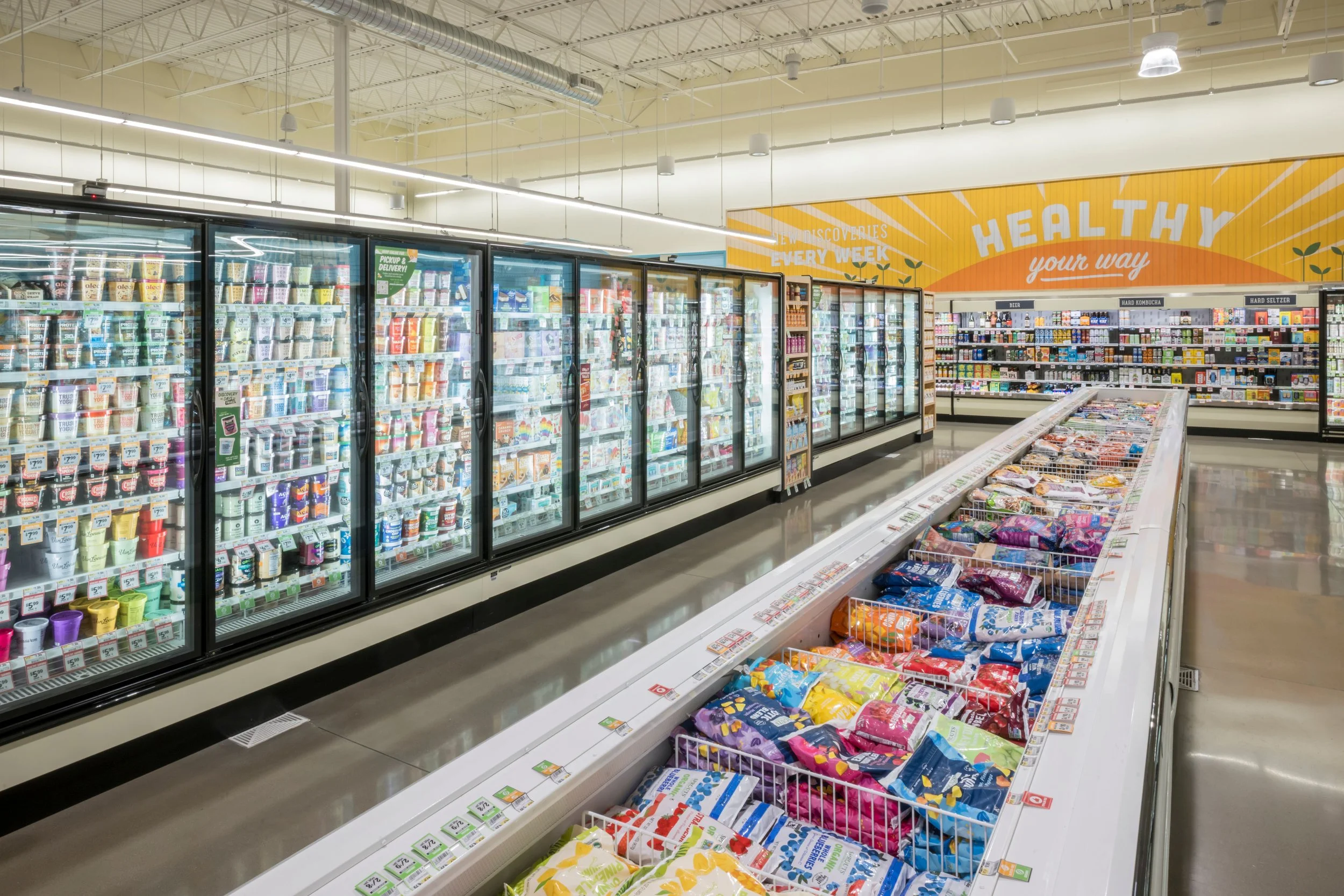 A grocery store aisle featuring ice cream in a freezer section, with shelves of snacks and candy in the background, and a brightly colored sign reading 'Healthy your way' on the wall.