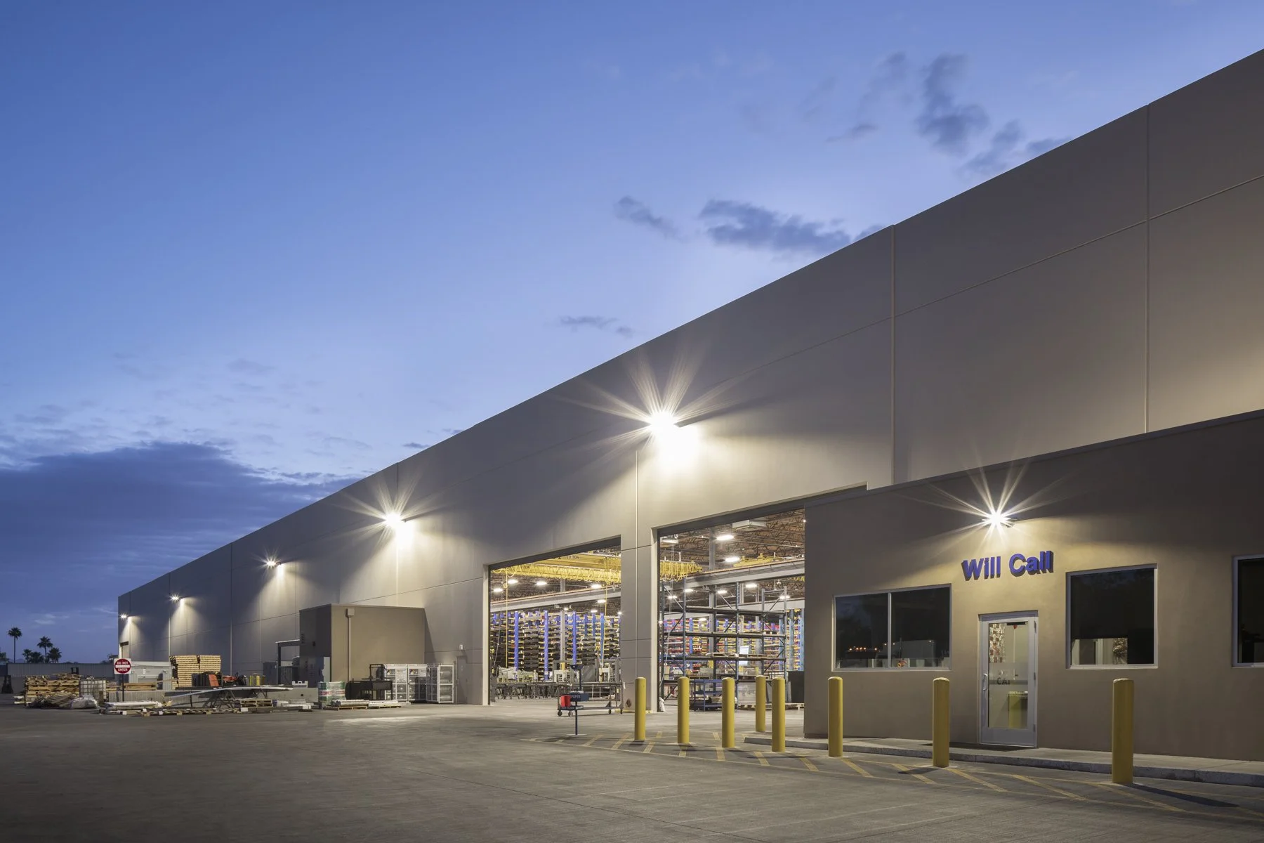 Exterior view of a large warehouse during dusk with lights illuminating the building and open garage door revealing shelves and industrial equipment inside. There are yellow bollards near the entrance labeled 'Will Call' and palm trees in the backgro