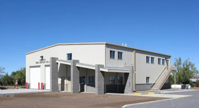 Exterior view of a two-story fire station building with a white facade, gray accents, and an outdoor staircase, under a clear blue sky.