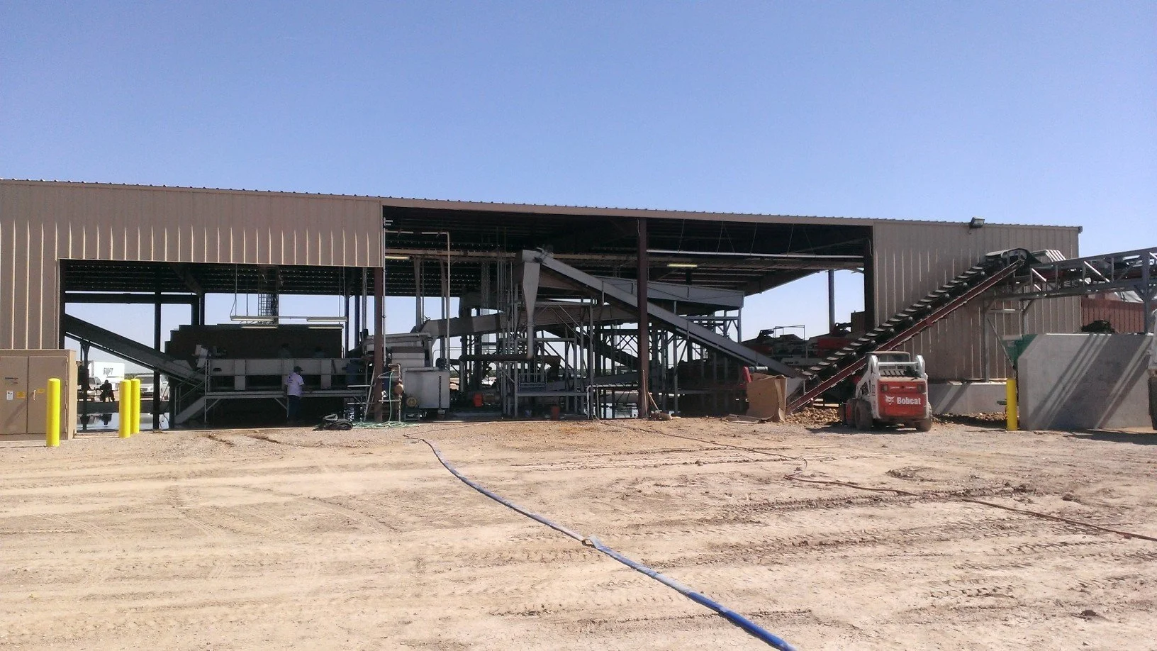 Industrial construction site with metal structure, conveyor belts, and a Bobcat skid-steer loader on dirt ground under clear blue sky.
