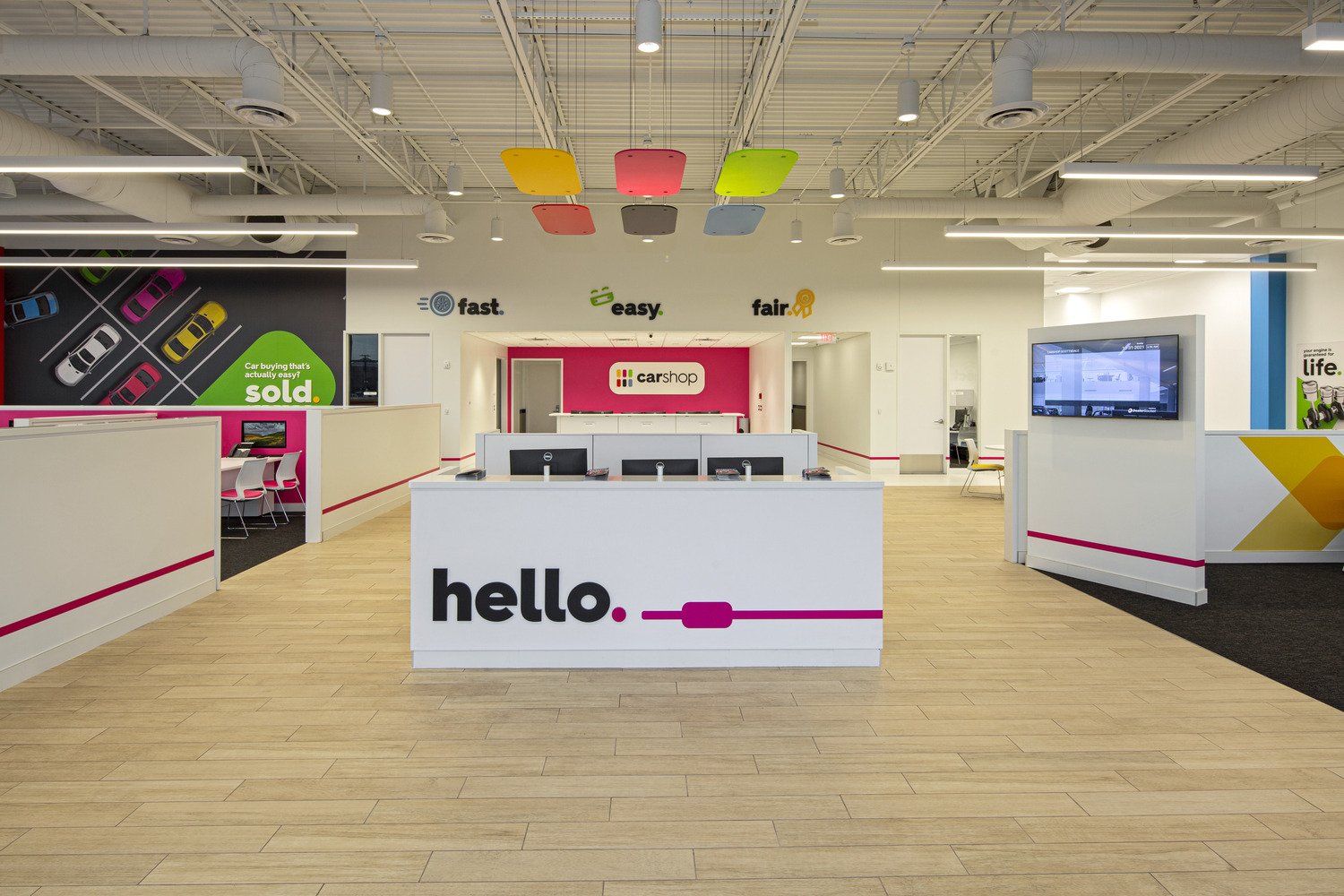 Interior of a modern car dealership reception area with a white front desk displaying the word 'hello' in large black letters and a pink dot and line design. Behind the desk are three computer monitors. The background features a wall with the words '