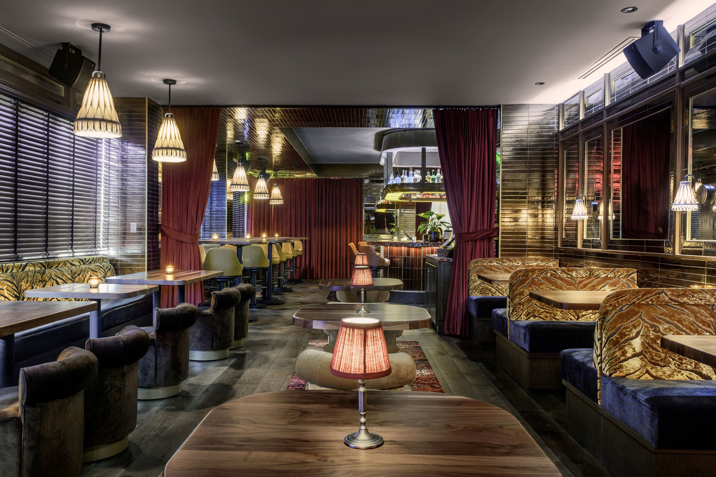 Interior of a cozy, upscale bar or restaurant with dark wood furniture, velvet seating, pendant lighting, and a bar area in the background.
