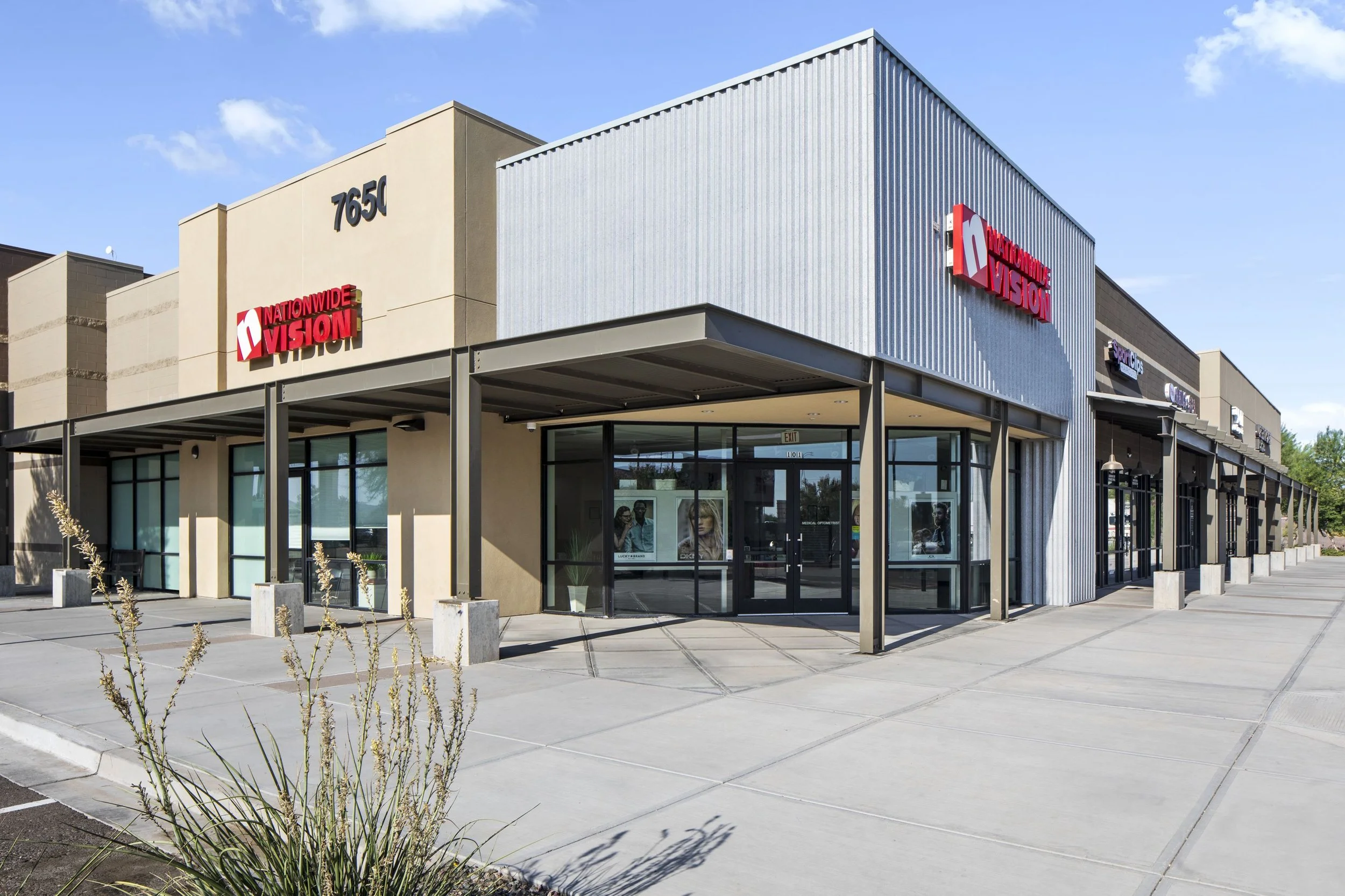Exterior of a retail store with a beige and gray facade, large windows, and red signage for 'Nationwide Vision' at 7650, with a parking lot in the foreground.