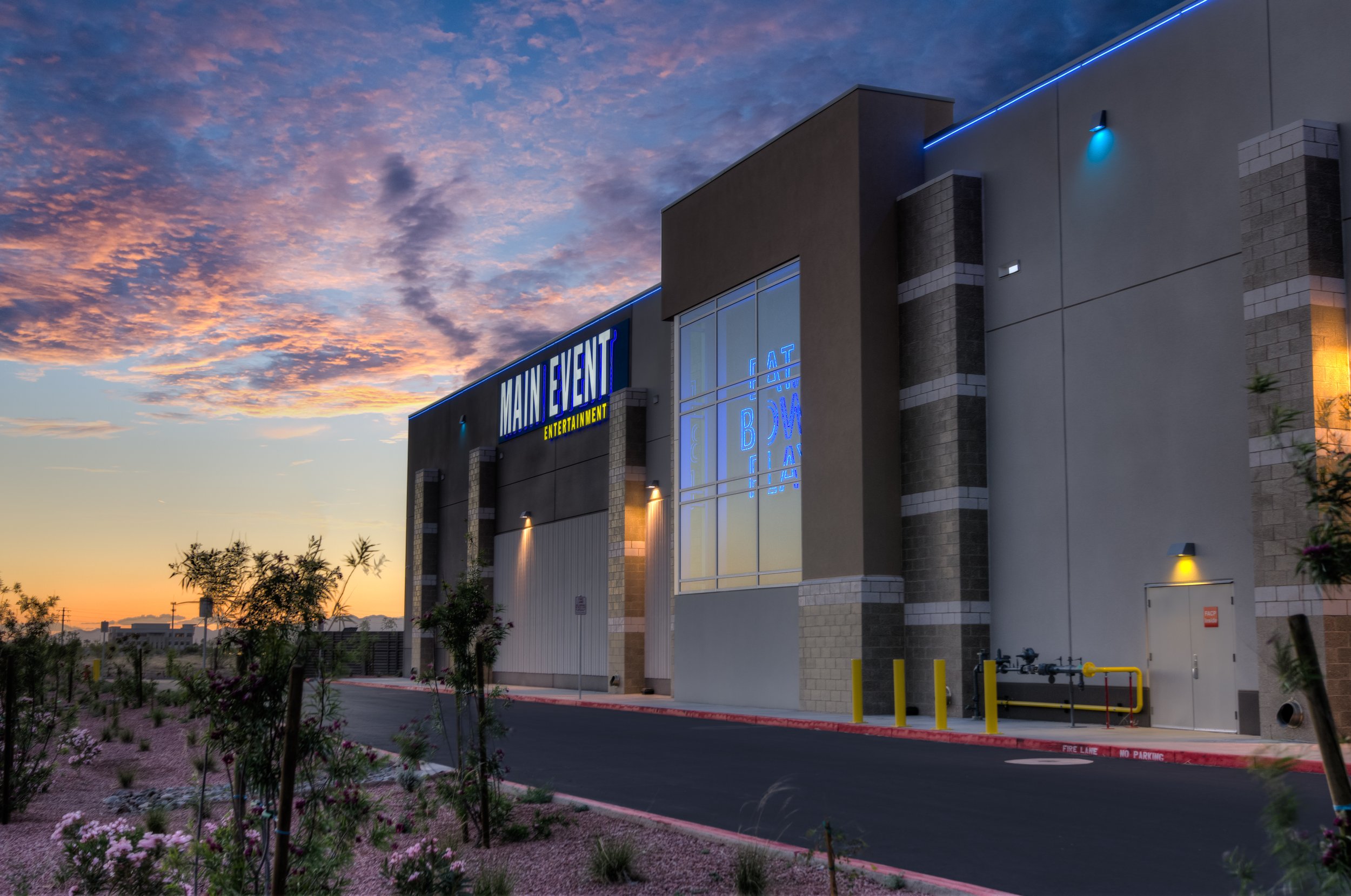 Exterior of a modern entertainment venue called Main Event at sunset, with illuminated signs, trees, and flowering plants in the foreground.