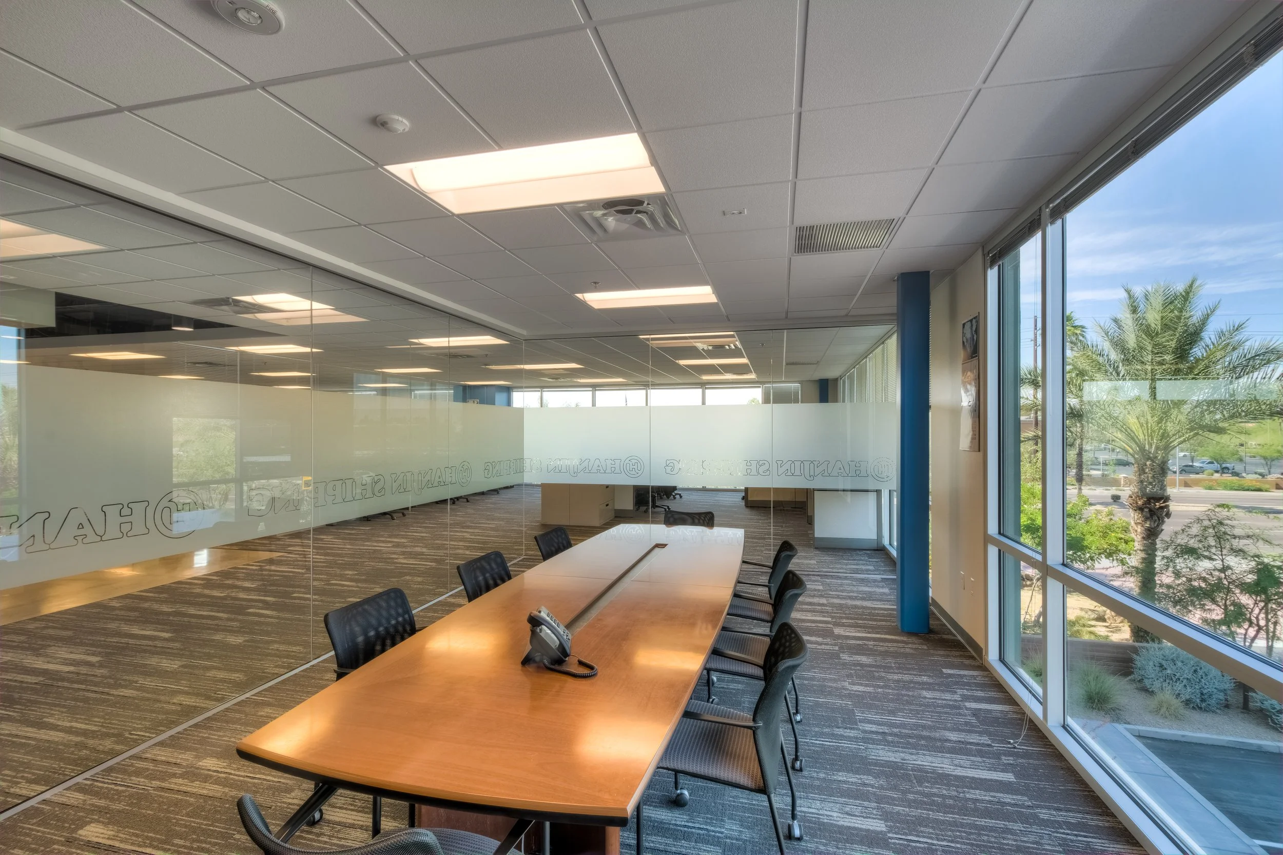 Empty conference room with a large wooden table, six chairs, a landline phone, and large windows showing palm trees outside.