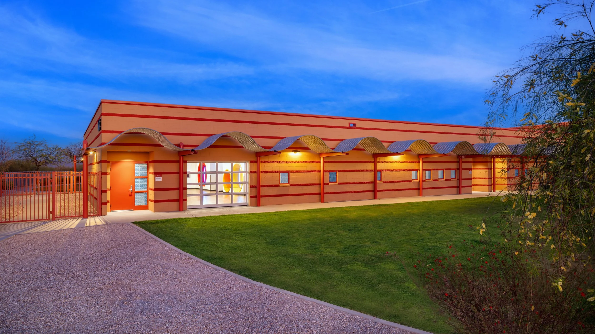 A modern building with a colorful, striped exterior and arched awnings, illuminated by exterior lights, with a well-manicured lawn and a gravel pathway in the foreground, during twilight.