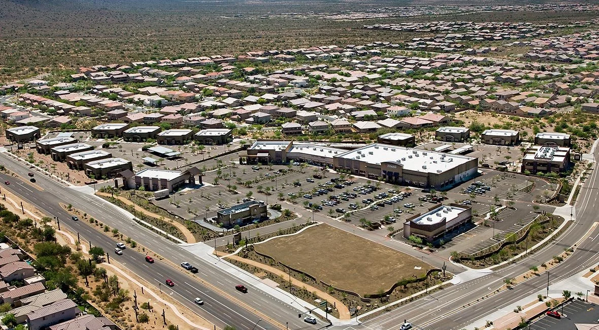 An aerial view of a shopping center with parking lots, surrounded by residential neighborhoods in a desert landscape.