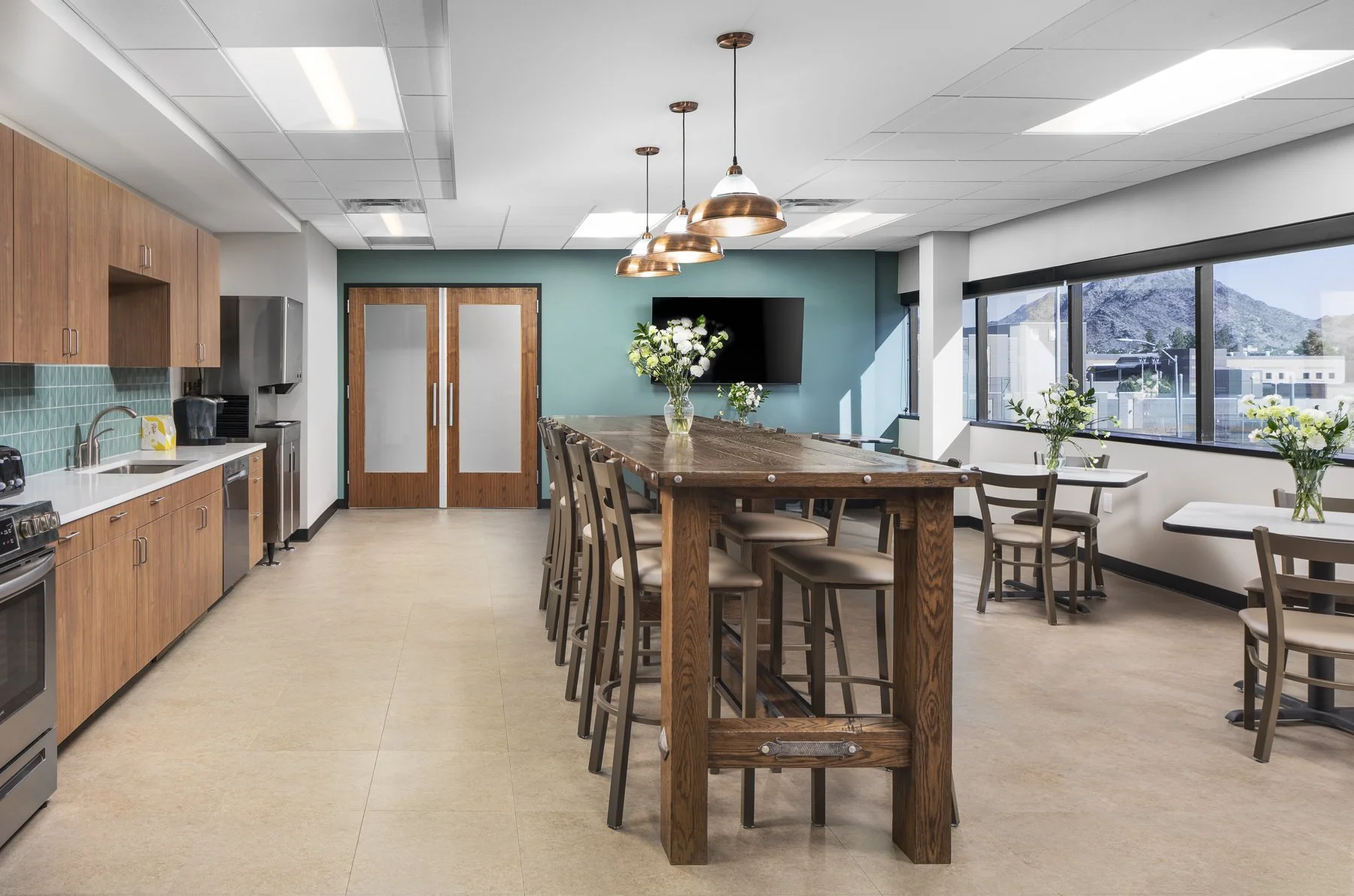 A modern cafeteria with a wooden high-top table surrounded by stools, painted green accent wall with a TV, large windows showing mountains outside, and a kitchenette with brown cabinets and green tiled backsplash.