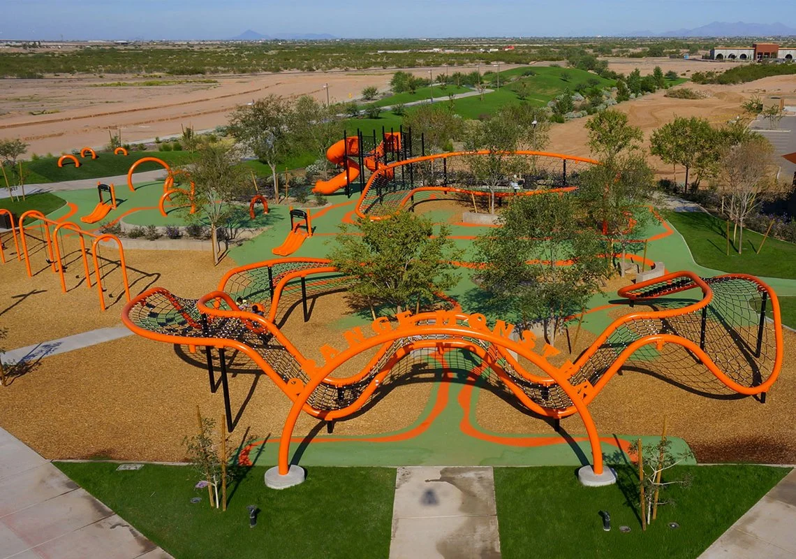 Colorful outdoor playground with orange and green equipment, including slides and climbing structures, in a park with trees and pathway, in a suburban area.
