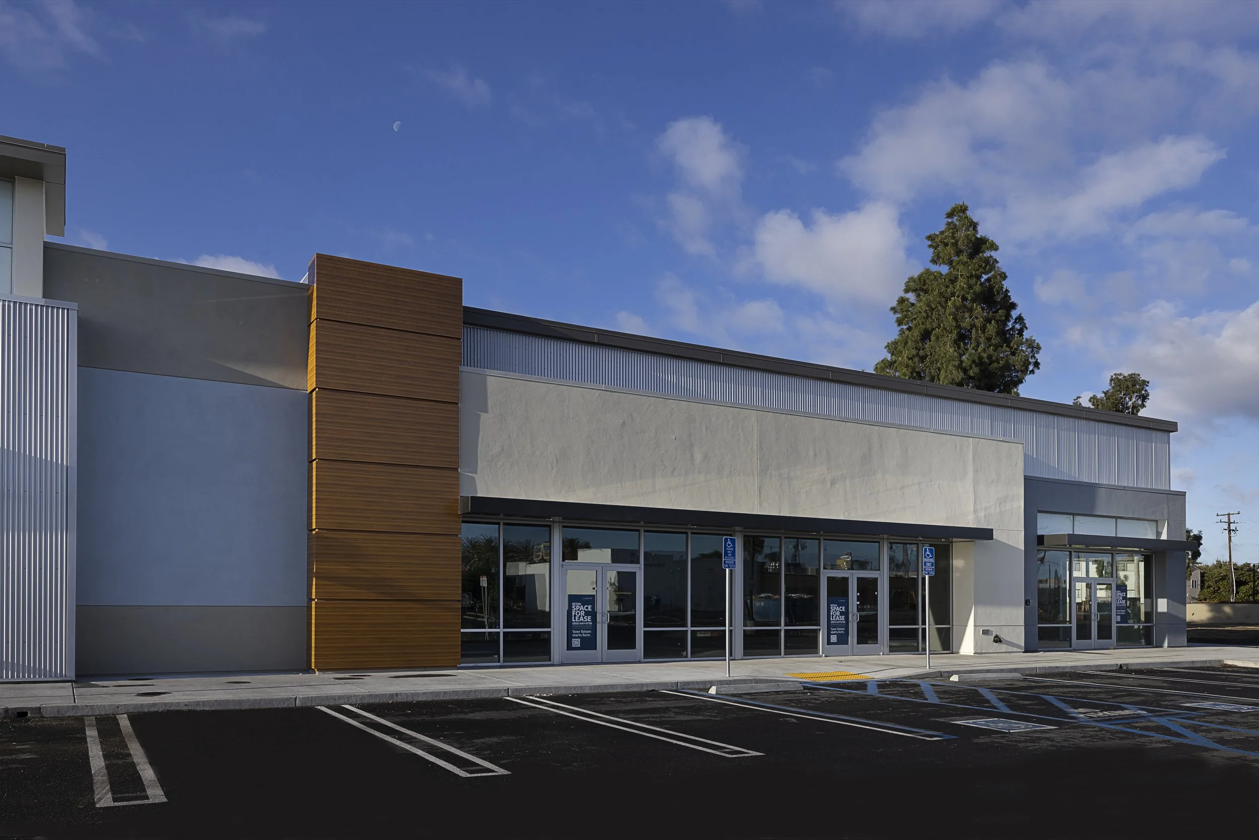 Modern commercial building with large glass windows and metal siding, with parking spaces in front and a parking sign, under a partly cloudy blue sky with a visible moon and a tree to the right.