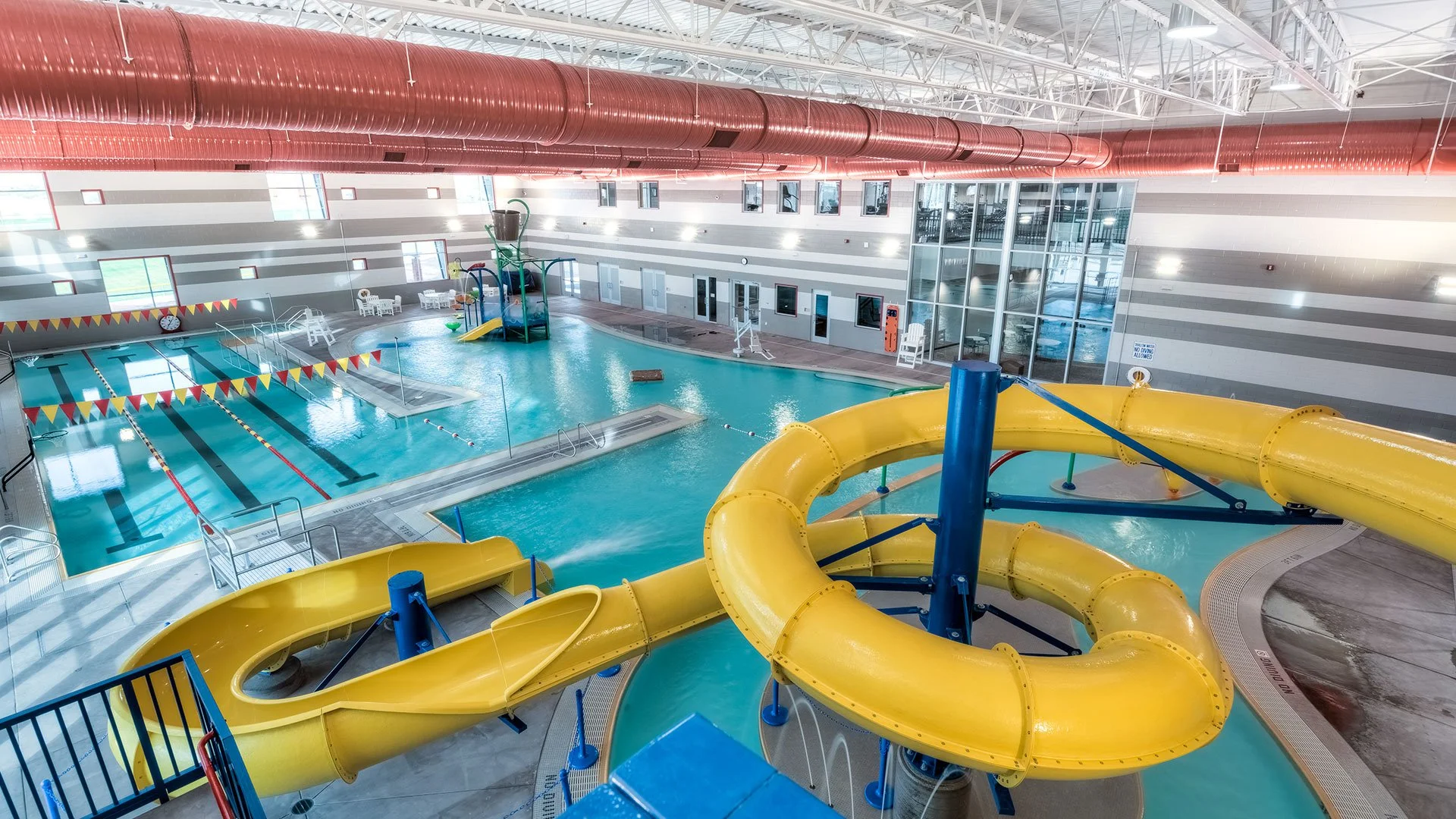 Indoor swimming pool area with water slides, a lazy river, and a children's play structure, surrounded by tiled floors and glass walls, with bright lighting.