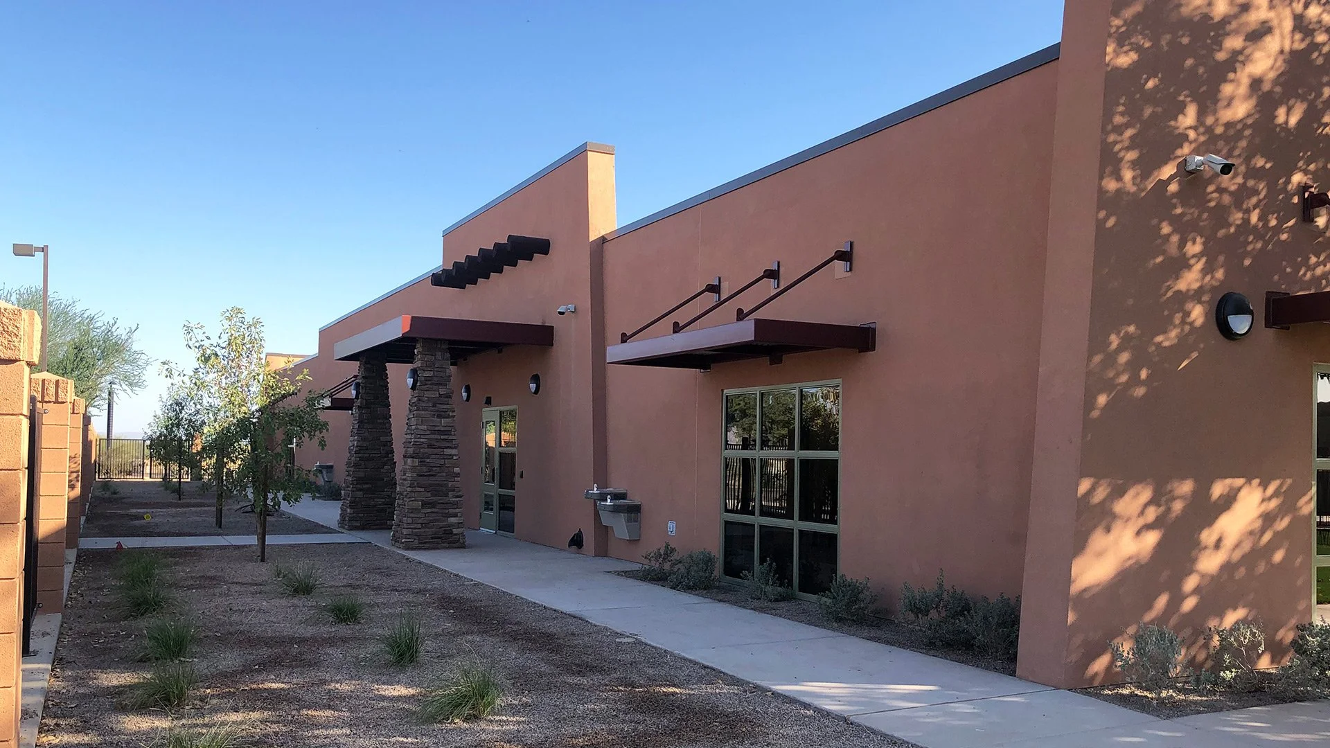 A modern building with a pink stucco exterior, large windows, and decorative awnings. The building has stone columns supporting the roof overhangs. There is a sidewalk with small plants and a landscaped area with young trees. Shadows from nearby tree