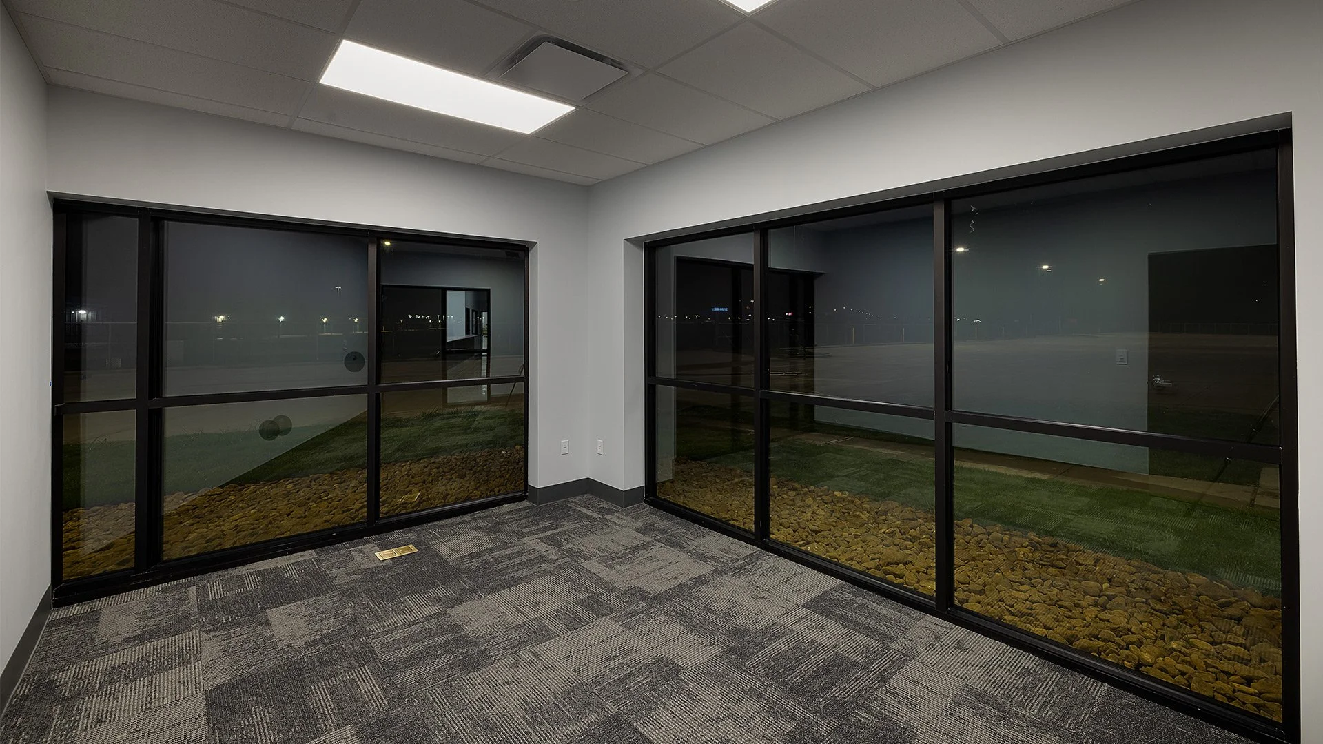 Empty office room with large glass windows showing an outdoor night view, carpeted floor, white walls, and ceiling with fluorescent lights.