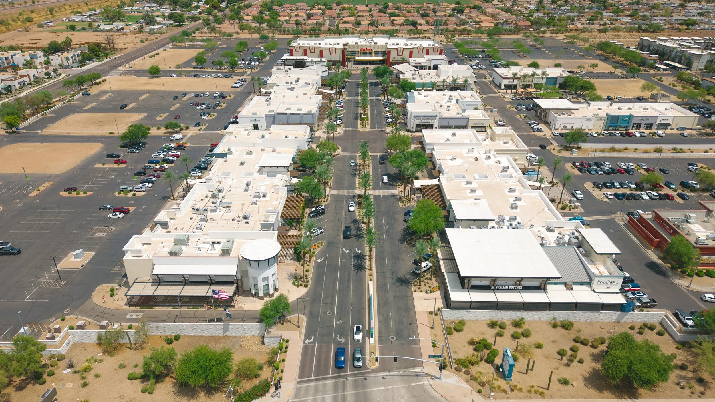 Aerial view of a shopping center with white buildings, parking lots, trees, and a large Harkins movie theater at the back. Surrounding areas include residential neighborhoods and undeveloped land.