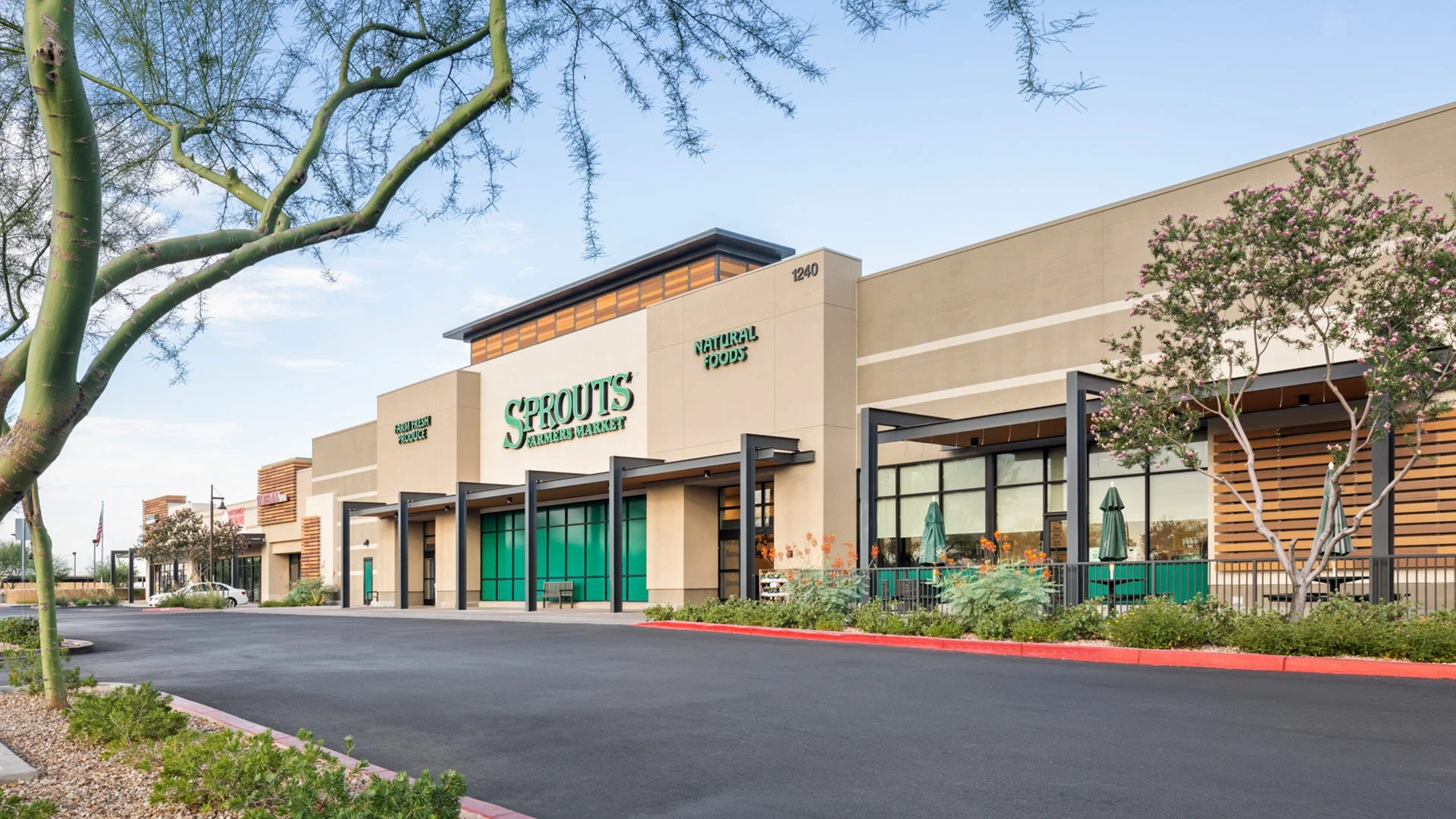 Exterior of a Sprouts Farmers Market grocery store in a shopping plaza, featuring a beige facade with green signage, outside seating area with umbrellas, trees, and a parking lot.