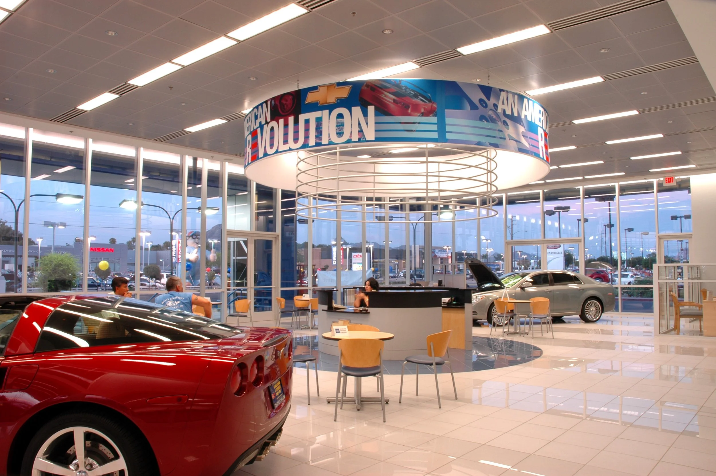 Inside an car dealership showroom with several cars on display, including a red sports car in the foreground, a silver sedan with open hood in the background, and a few customers and staff at a central counter. The showroom has large glass windows re