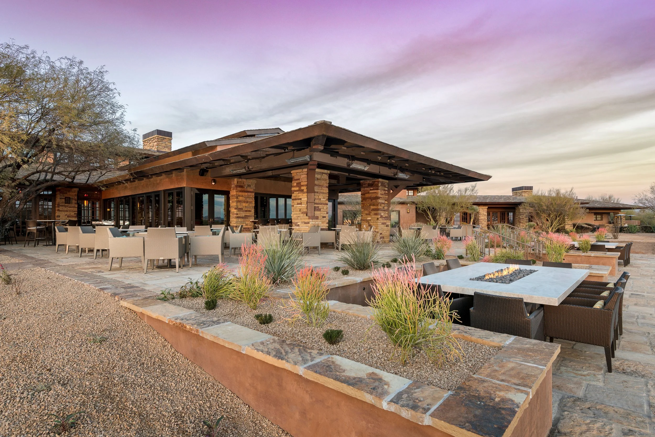 An outdoor patio area of a restaurant with tables and chairs, a fire pit, and desert landscaping, under a sunset sky.
