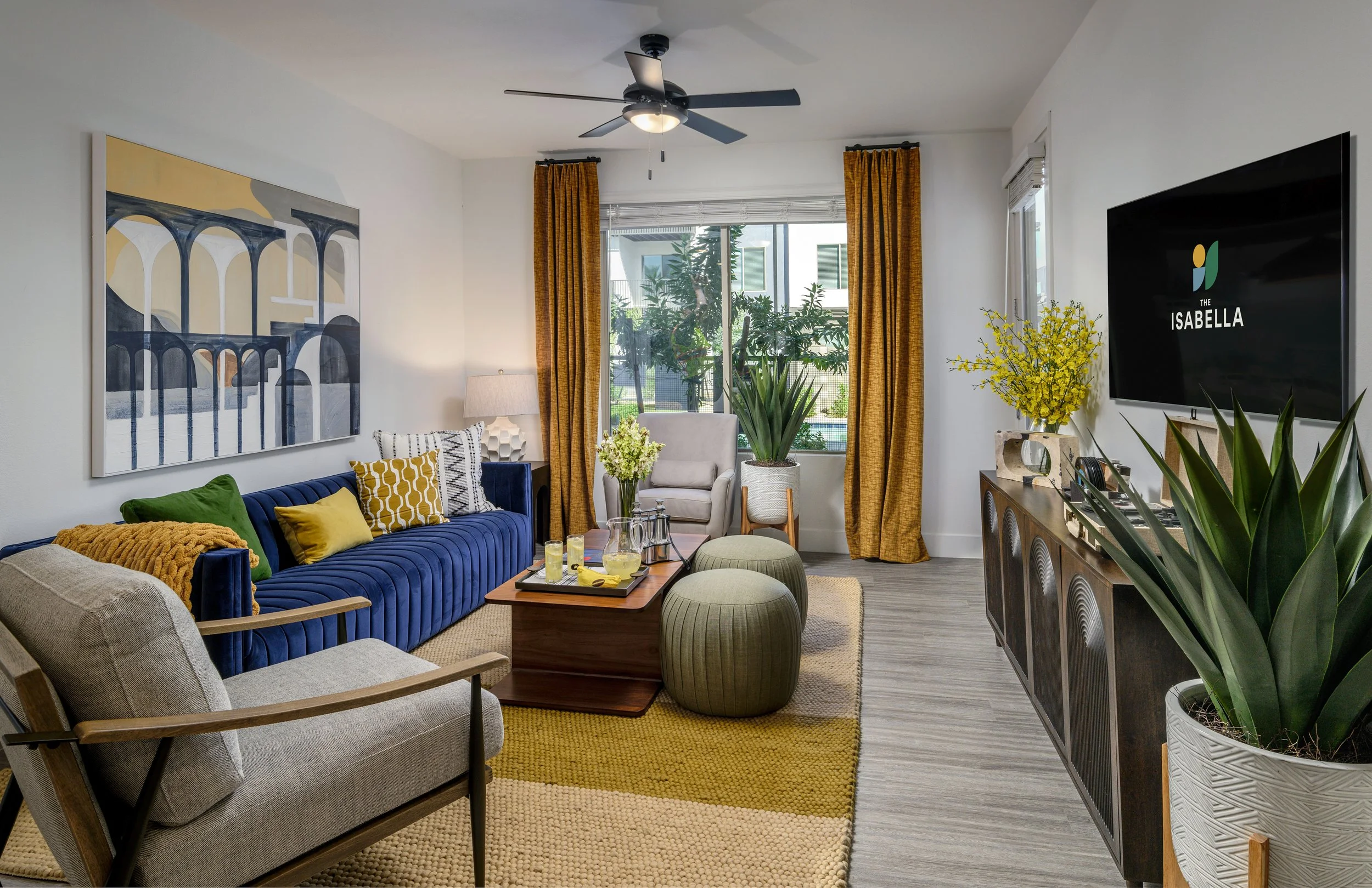 A modern living room with a blue sofa, armchair, and two green ottomans centered around a wooden coffee table with drinks and a tray. There's a large window with yellow curtains, potted plants, artworks, a wall-mounted TV, and a dark wood sideboard. 