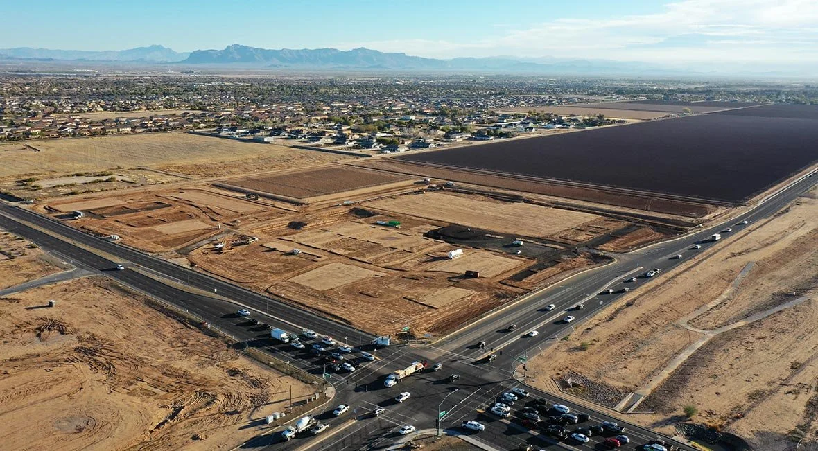 An aerial view of a construction site next to a busy road, with a city and mountains in the background.
