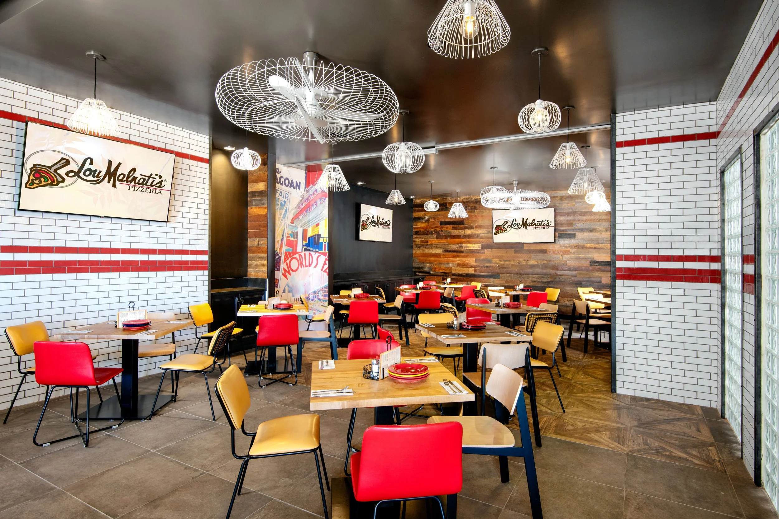 Interior of a pizzeria with wooden and brick walls, multiple tables with colorful chairs, modern pendant lighting, and Lou Malnati's logo on the wall.