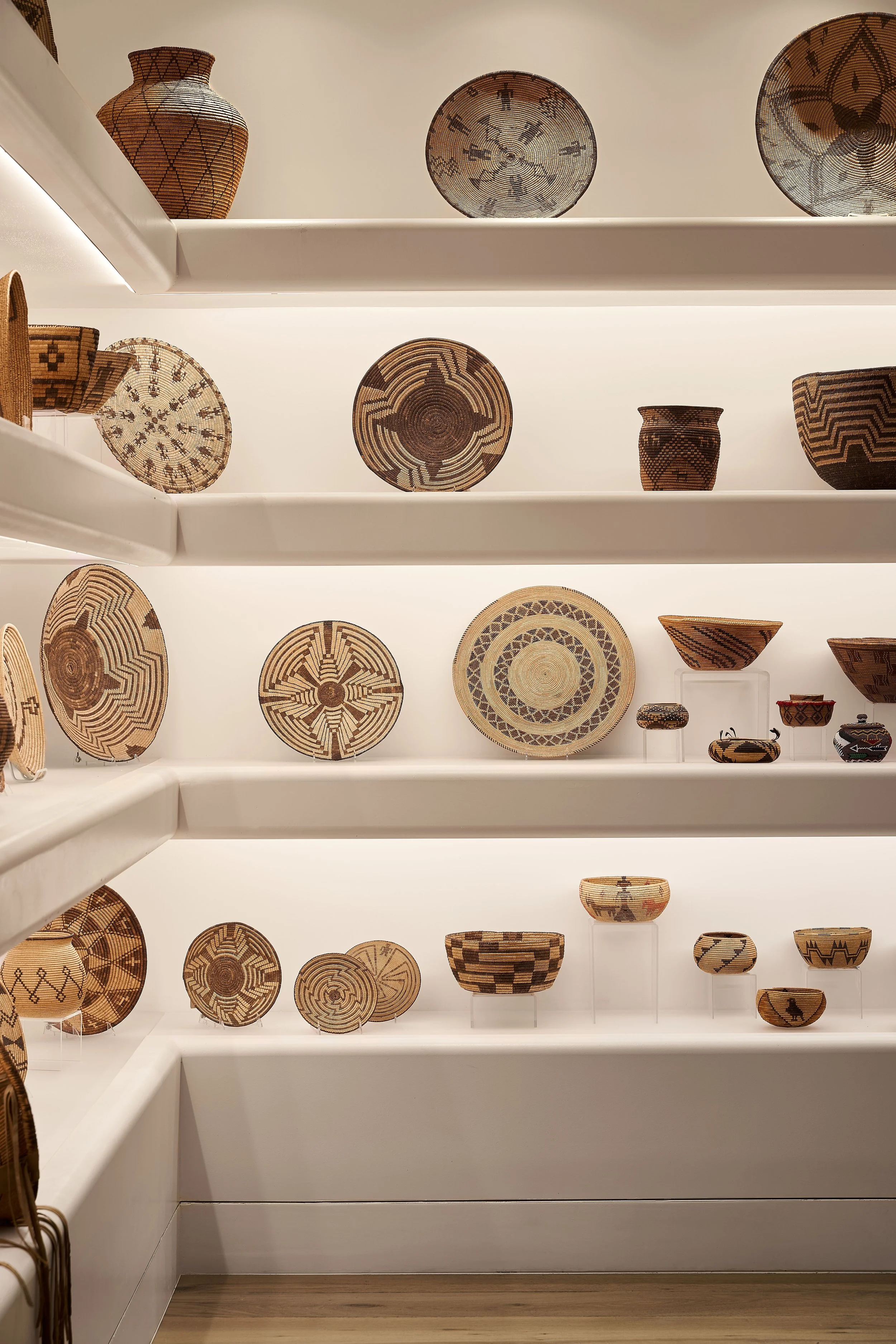 Display of woven baskets and bowls on white shelves in a museum or gallery setting.