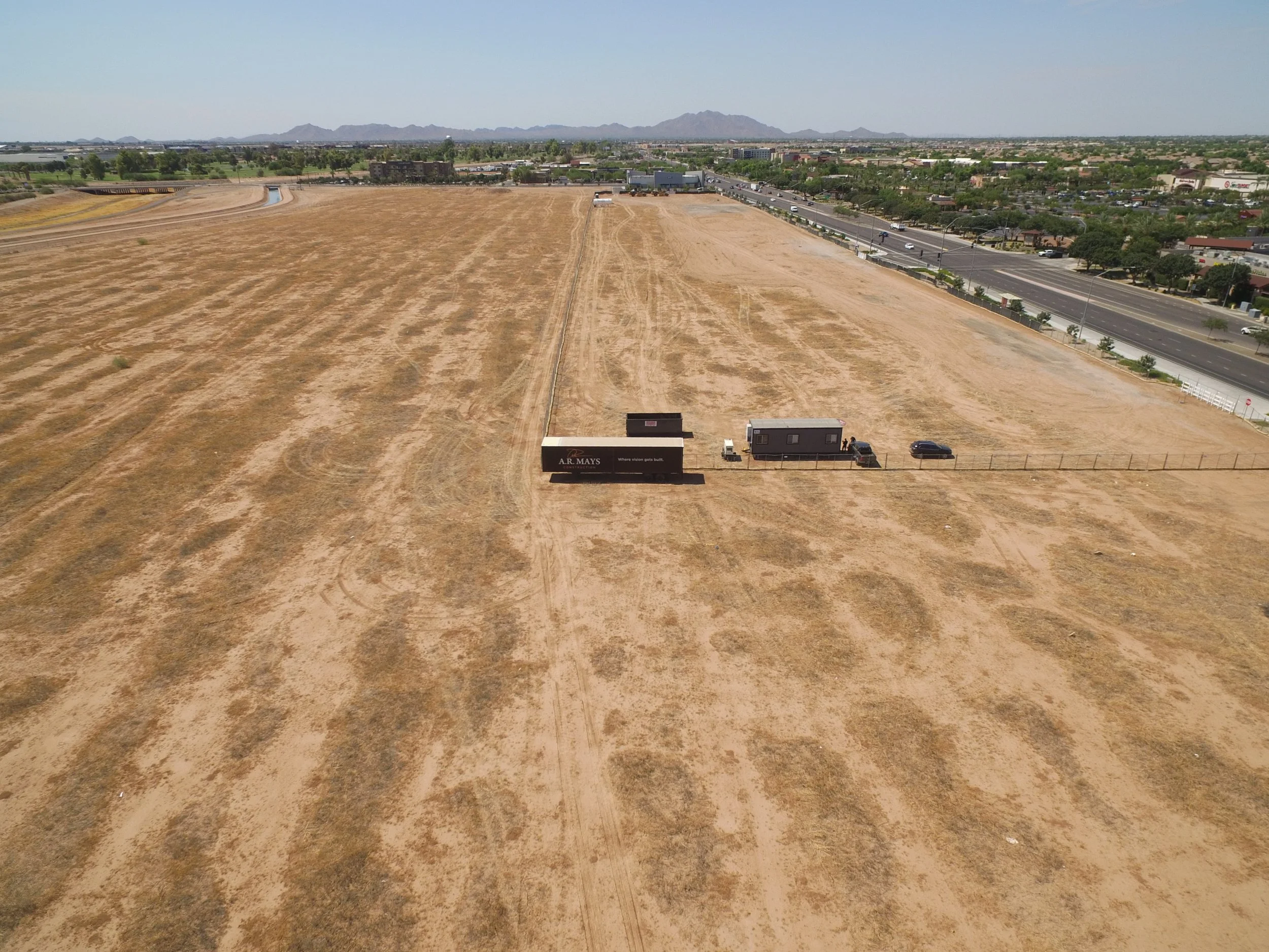 Empty dry field with two trailers, one black and one gray, and two cars parked near a fence. City roads and buildings in the background, with mountains in the distance.