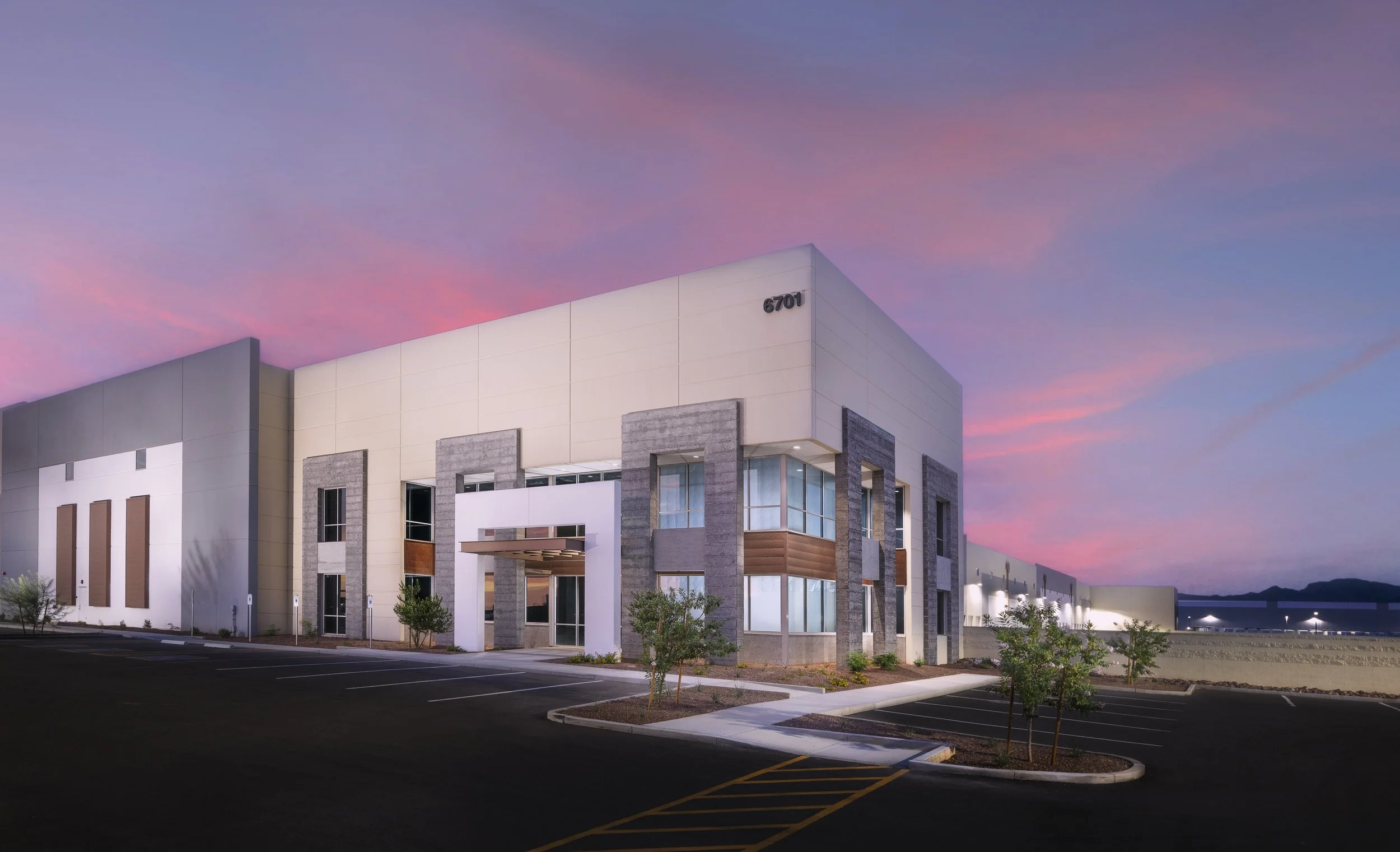 Modern commercial building with large windows and neutral colors during sunset, with a parking lot in front and trees around