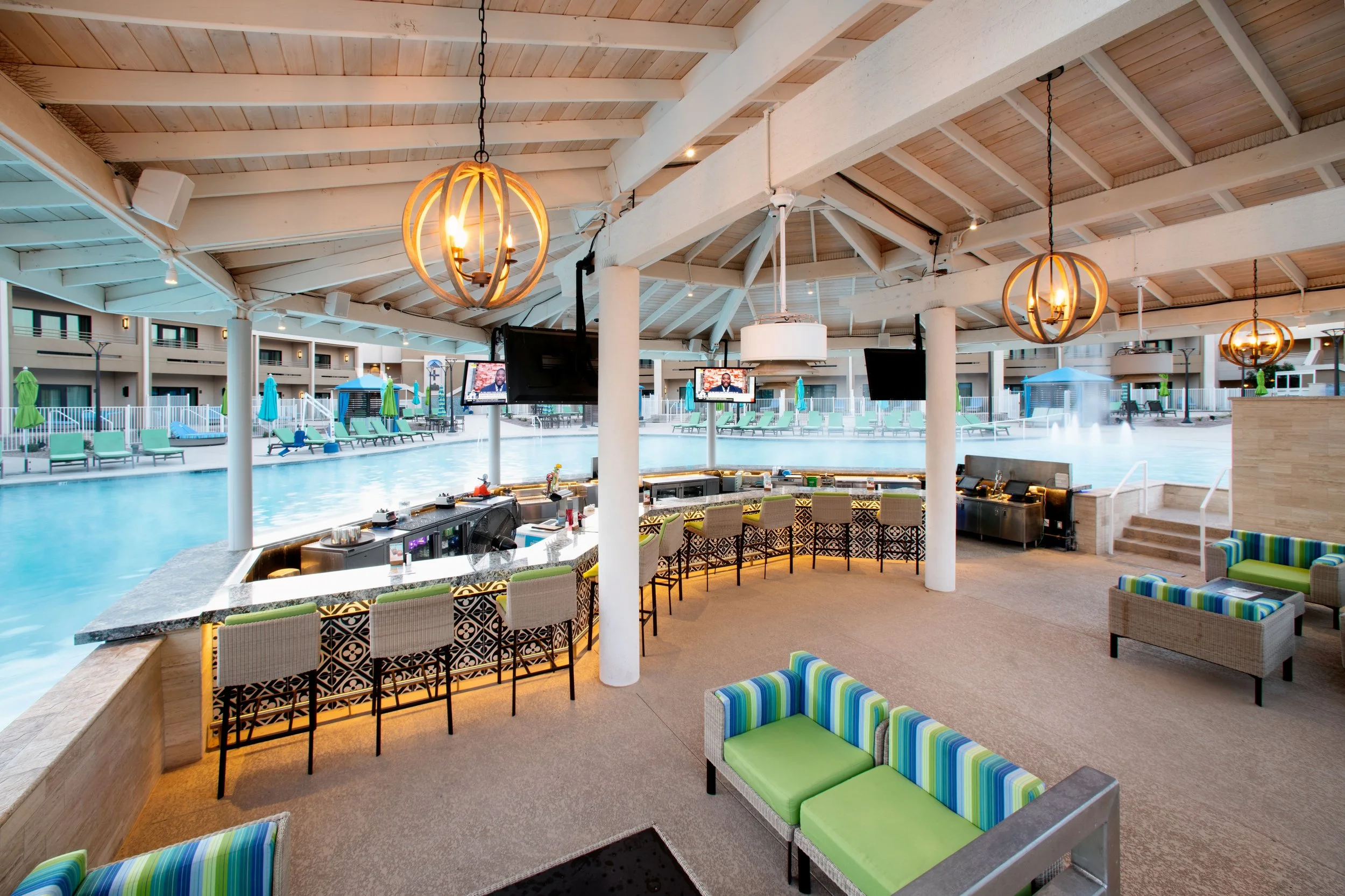 Indoor pool area with swim-up bar, bar stools, wall-mounted TVs, lounge chairs, and ceiling fans with warm lighting.