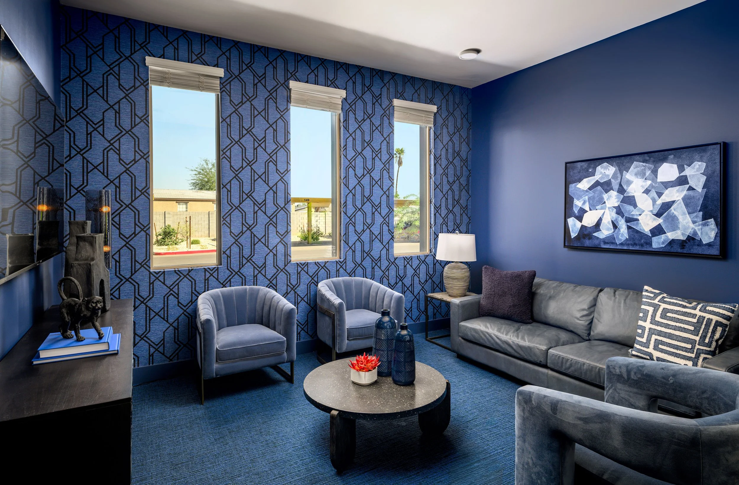 Living room with three windows, blue patterned accent wall, grey leather sofa, blue armchairs, a black coffee table with vases, and modern art
