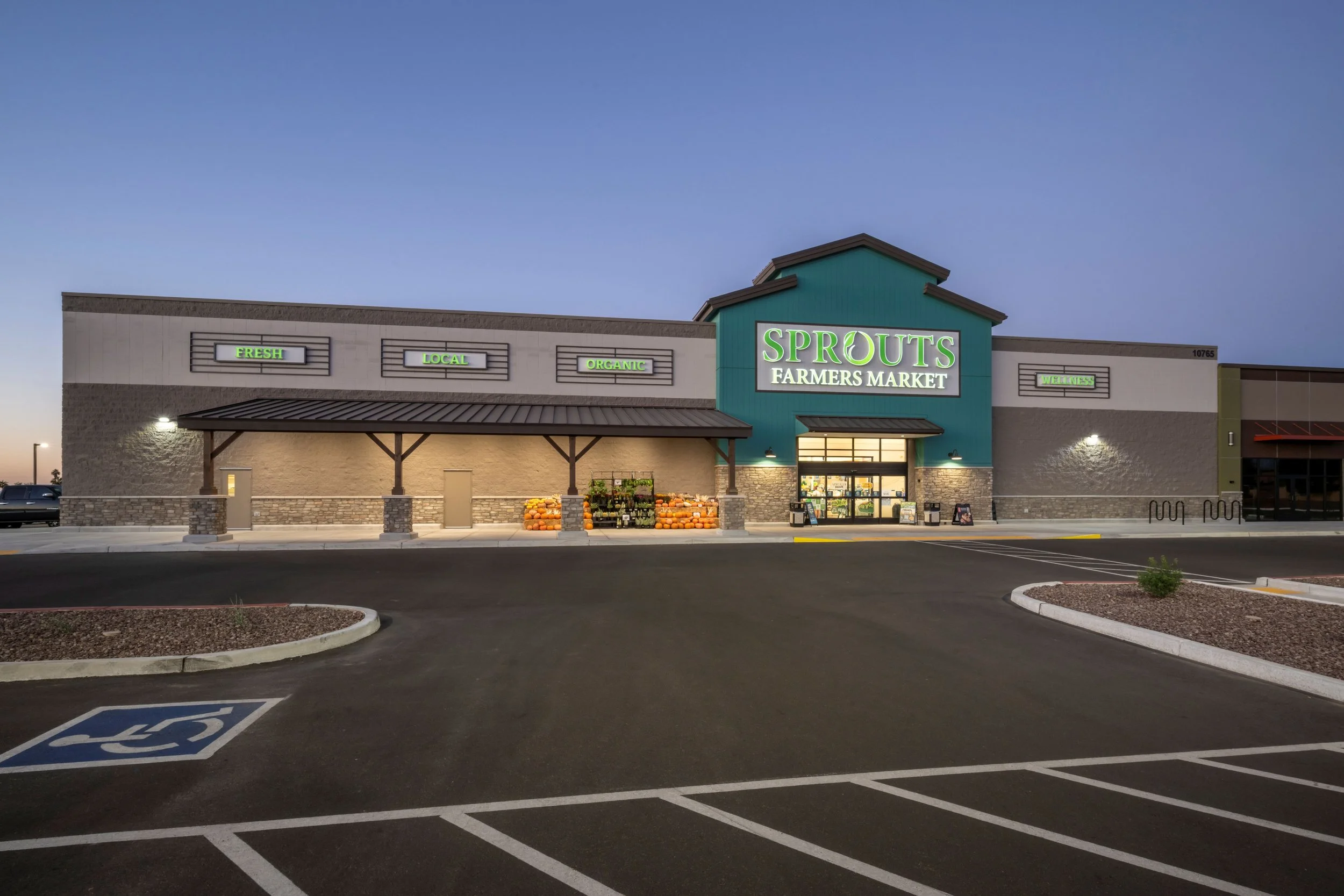 Exterior of Sprouts Farmers Market store during dusk with parking lot in foreground, including a handicapped parking space, and the store's sign illuminated.
