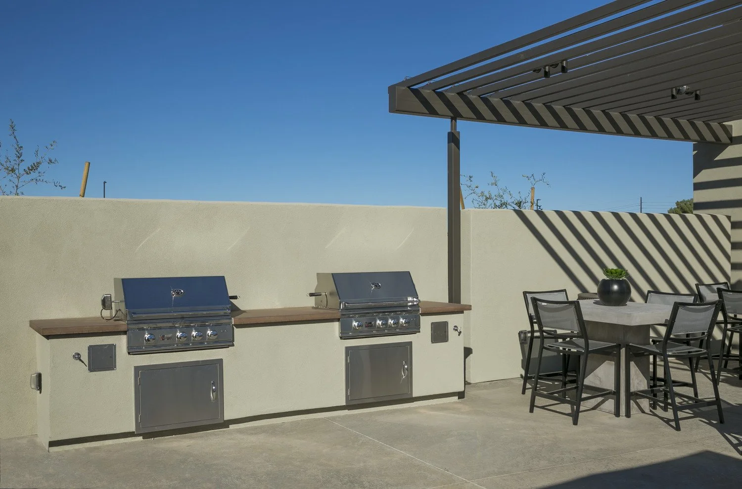 Outdoor patio area with two stainless steel gas grills, a black table with six chairs, a black planter with green plant, and a beige wall with a blue sky background.
