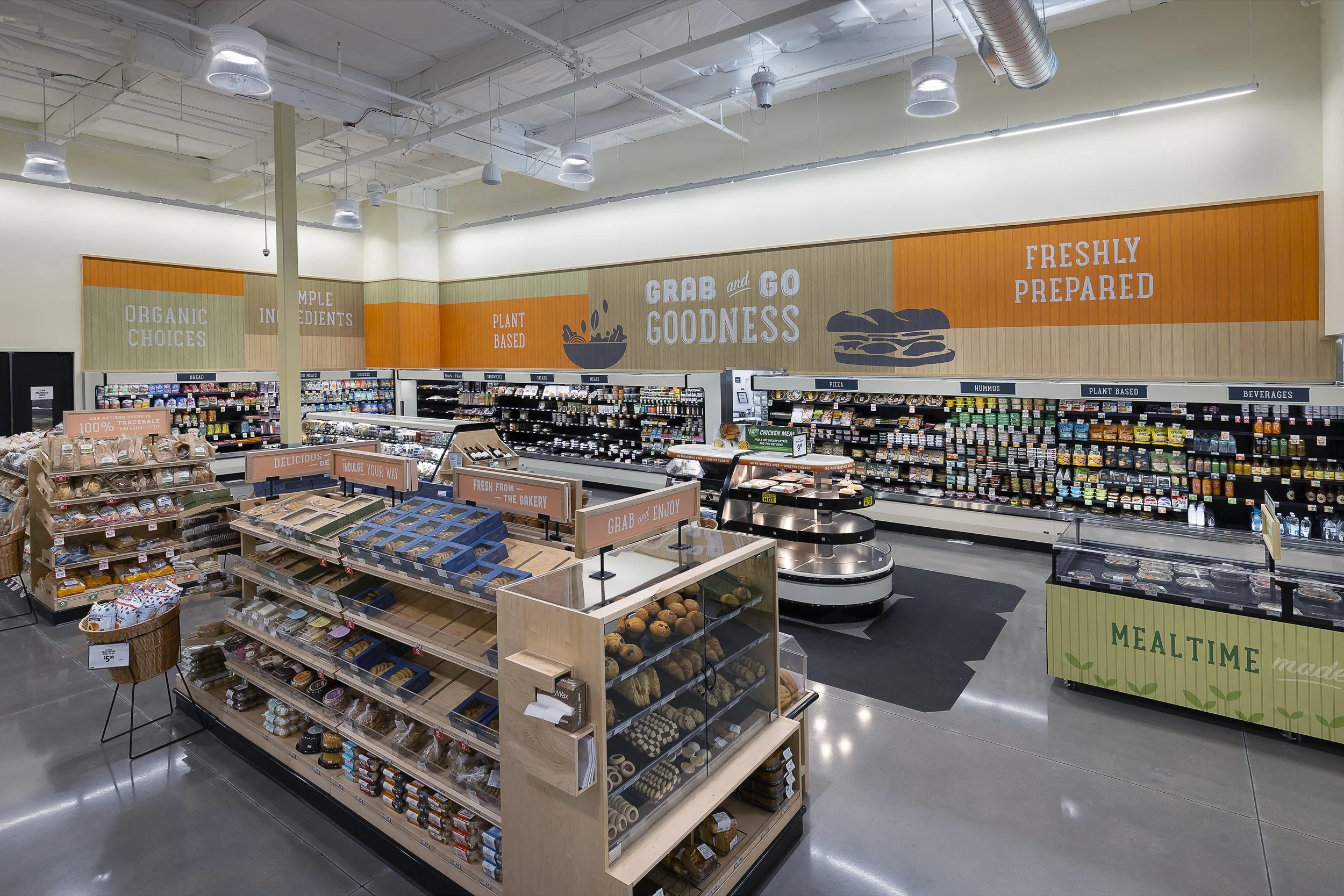 Interior view of a grocery store aisle with a bakery section and refrigerated goods on the shelves. Bright lighting and a modern design with signs highlighting organic choices, plant-based ingredients, and fresh, prepared foods.