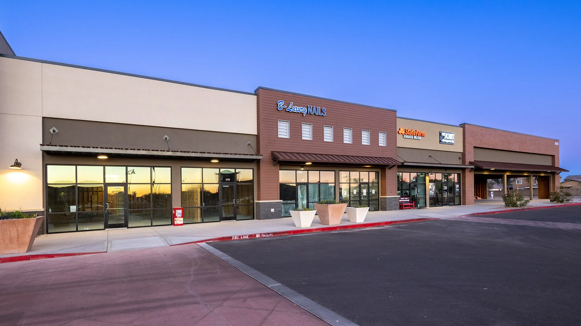Strip mall with various storefronts, including a nail salon, a State Farm insurance office, and other businesses, with a parking lot and sunset sky.