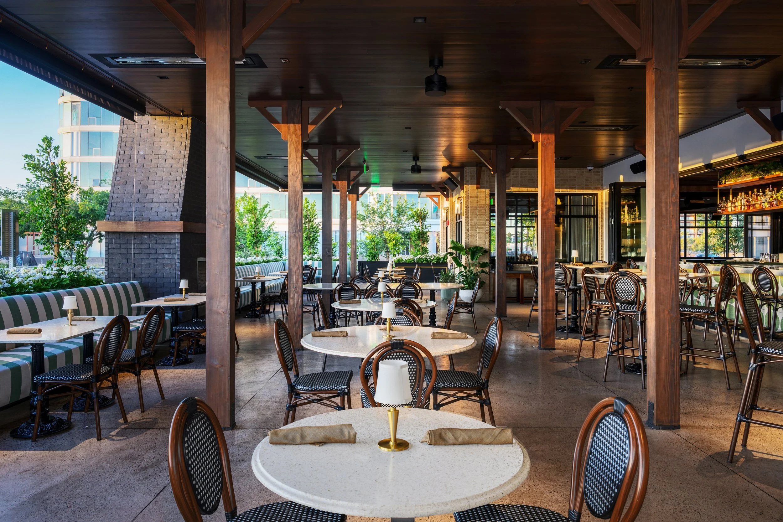 Empty restaurant with round tables, chairs, and bar area, natural light through large windows, decorated with plants.