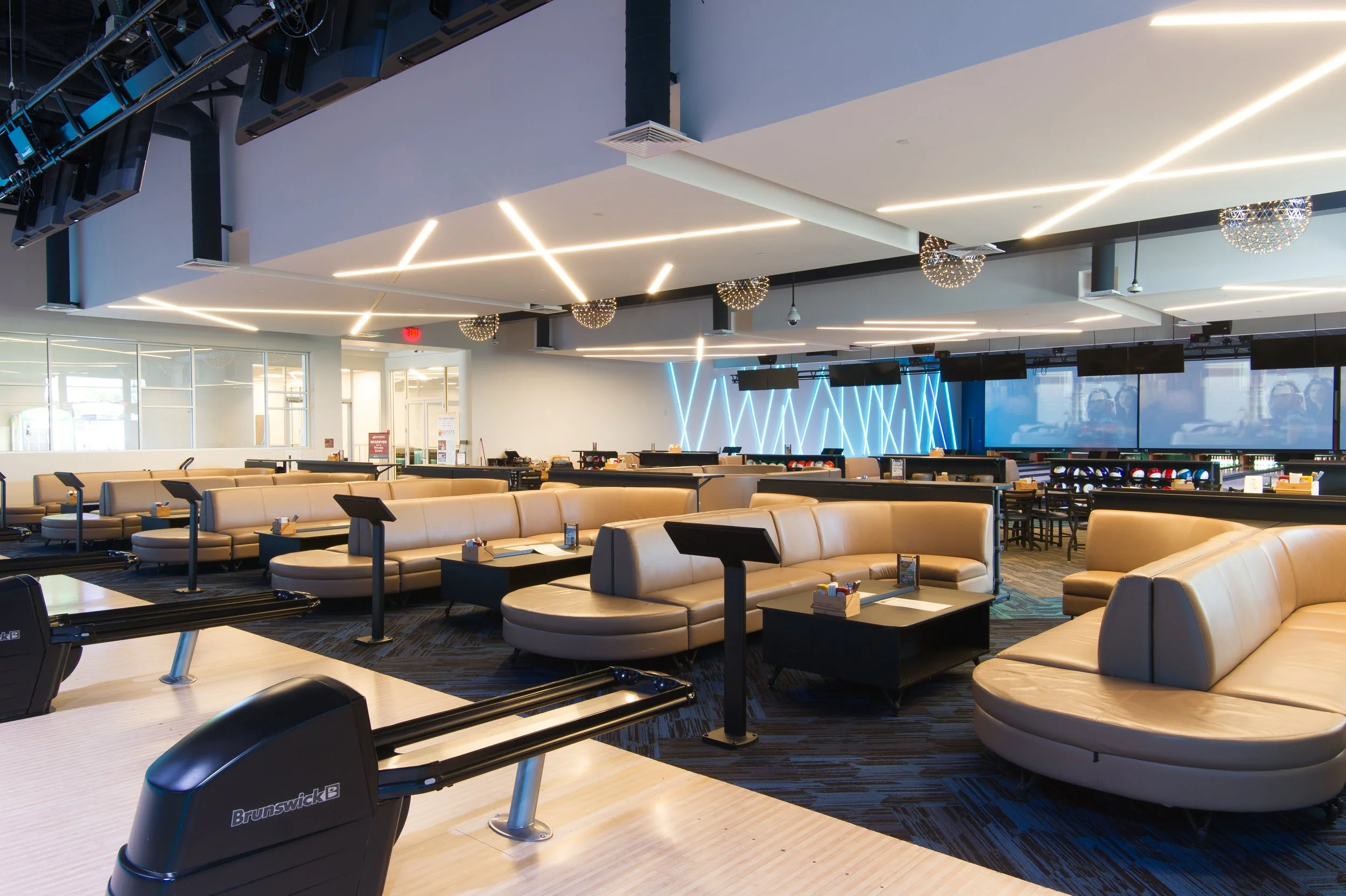 Interior of a modern, empty lounge or waiting area with beige curved sofas, small black tables, and a dark patterned carpet. The ceiling has geometric LED light fixtures and hanging spherical lights, with large windows and screens on one side.