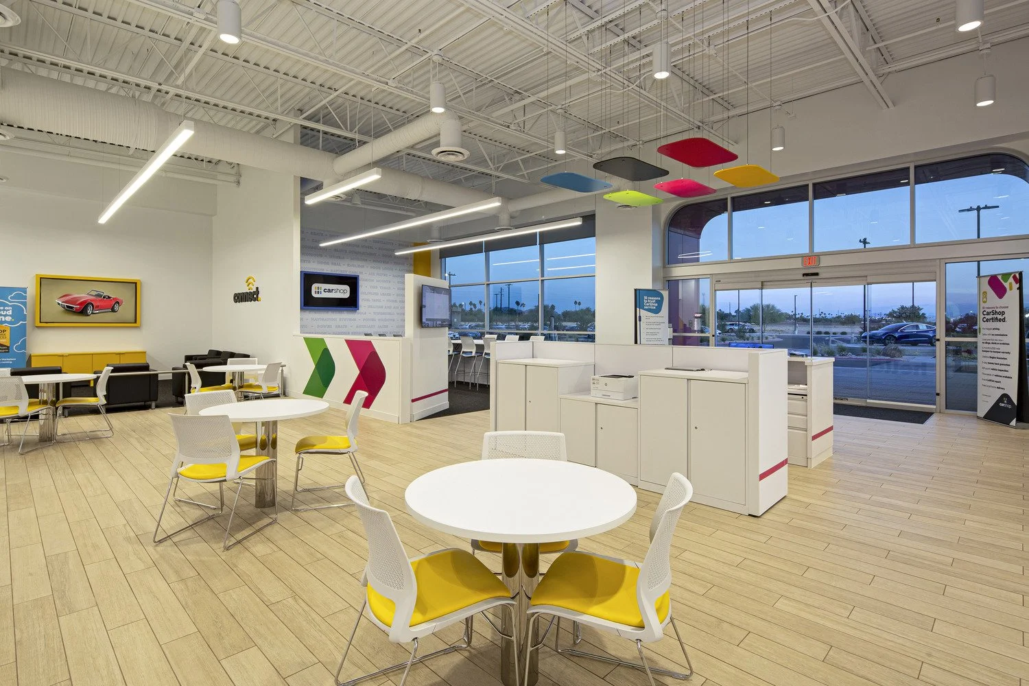 Interior of a modern car dealership waiting area with white walls, large windows, yellow and white chairs, round tables, and colorful ceiling panels.