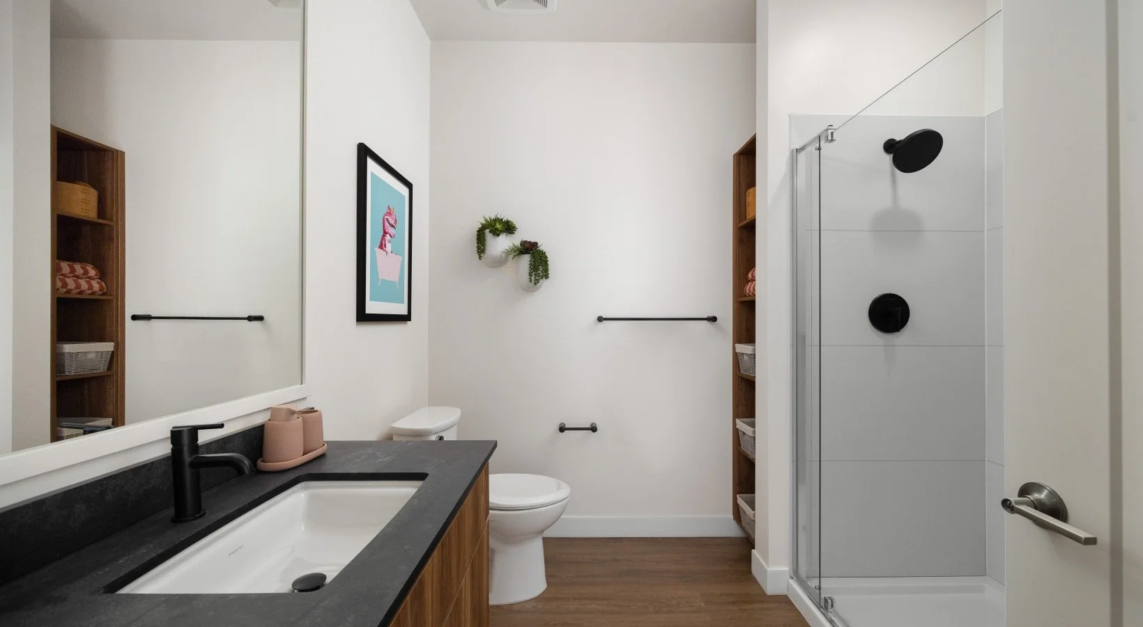A modern bathroom featuring a white toilet, a black countertop with a white sink and a black faucet, wooden shelves with baskets and towels, a large mirror, decorative wall hanging plants, and a glass shower with black fixtures.