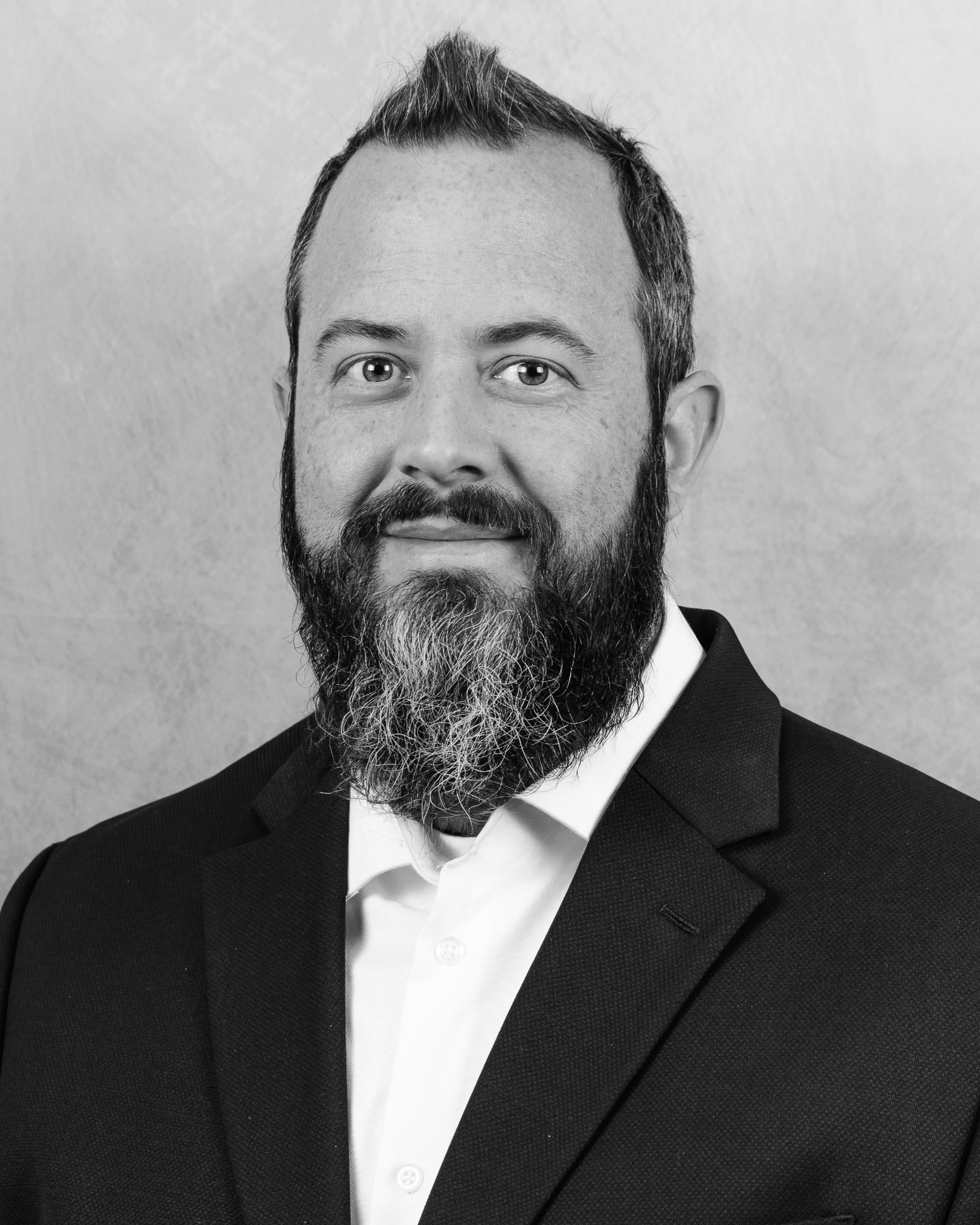 Black and white close-up photo of a smiling man with a beard and short hair, wearing a tuxedo and a white shirt.